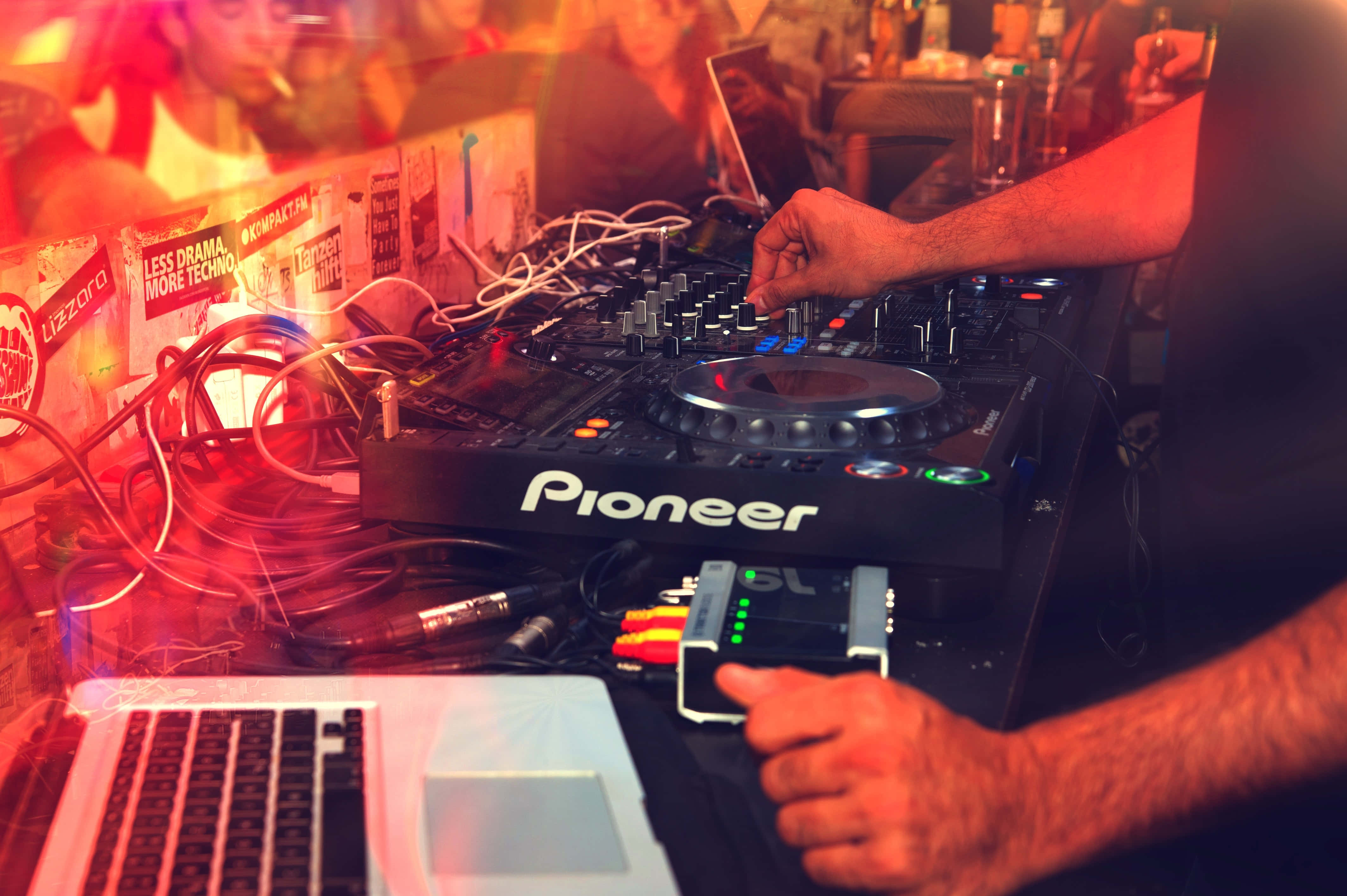 music festival 桌布 壁紙 — 一位DJ的手熟練地操作著Pioneer混音器，在充滿活力的紅色和橙色俱樂部燈光下，旁邊有纏繞的電線和一部筆記型電腦。