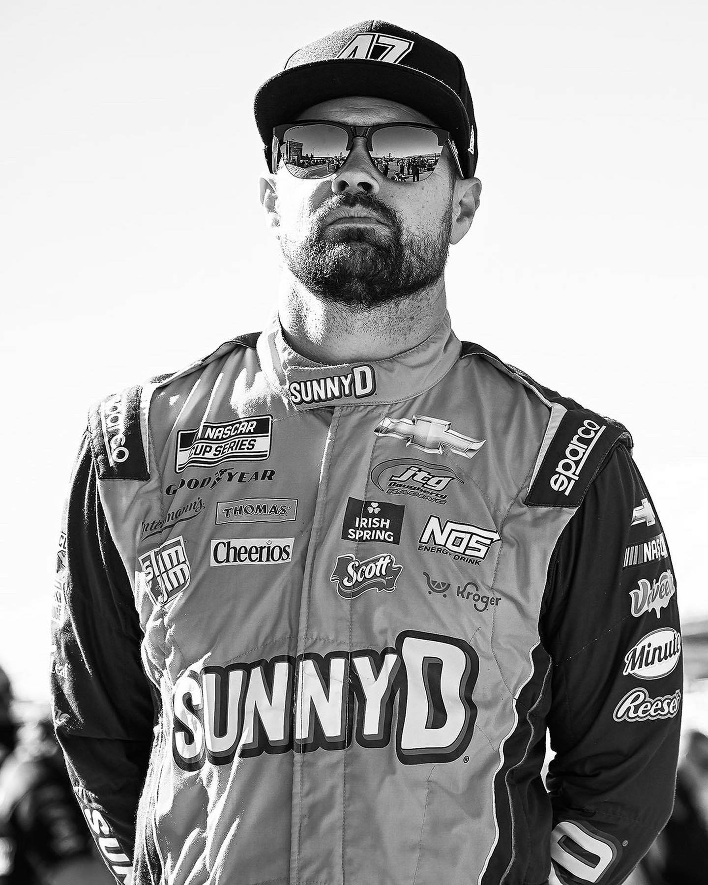 黑白 桌布 壁紙 — 一位NASCAR車手的黑白肖像，身穿贊助商覆蓋的賽車服和帽子，戴著反射人群的太陽眼鏡。