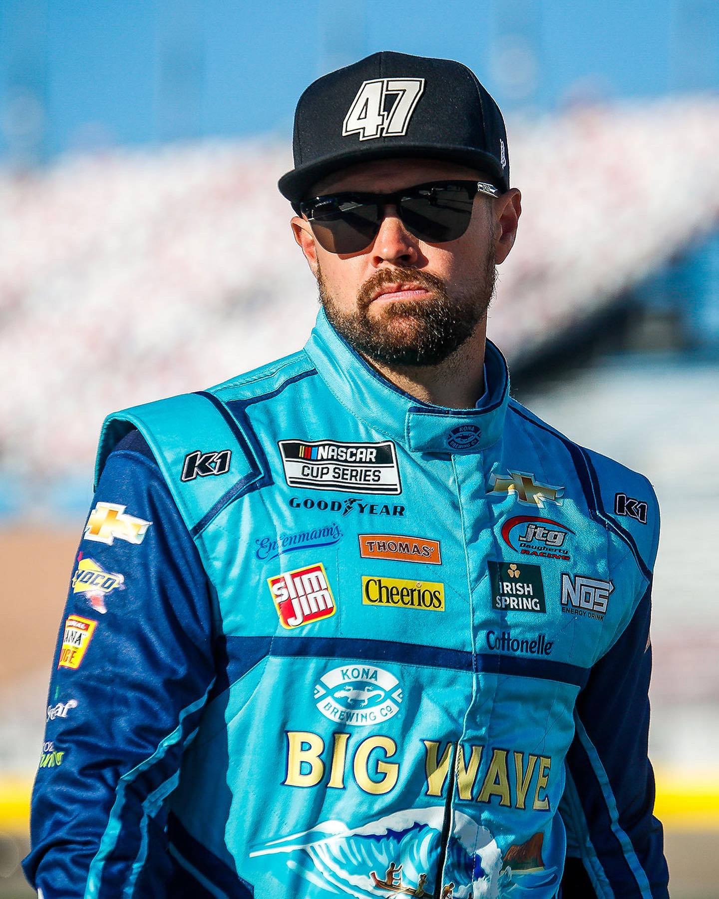 Athlete 桌布 壁紙 — 一位NASCAR車手穿著藍色和藍綠色的賽車服，帶有贊助商標誌，在明亮的日光下自信地站立。