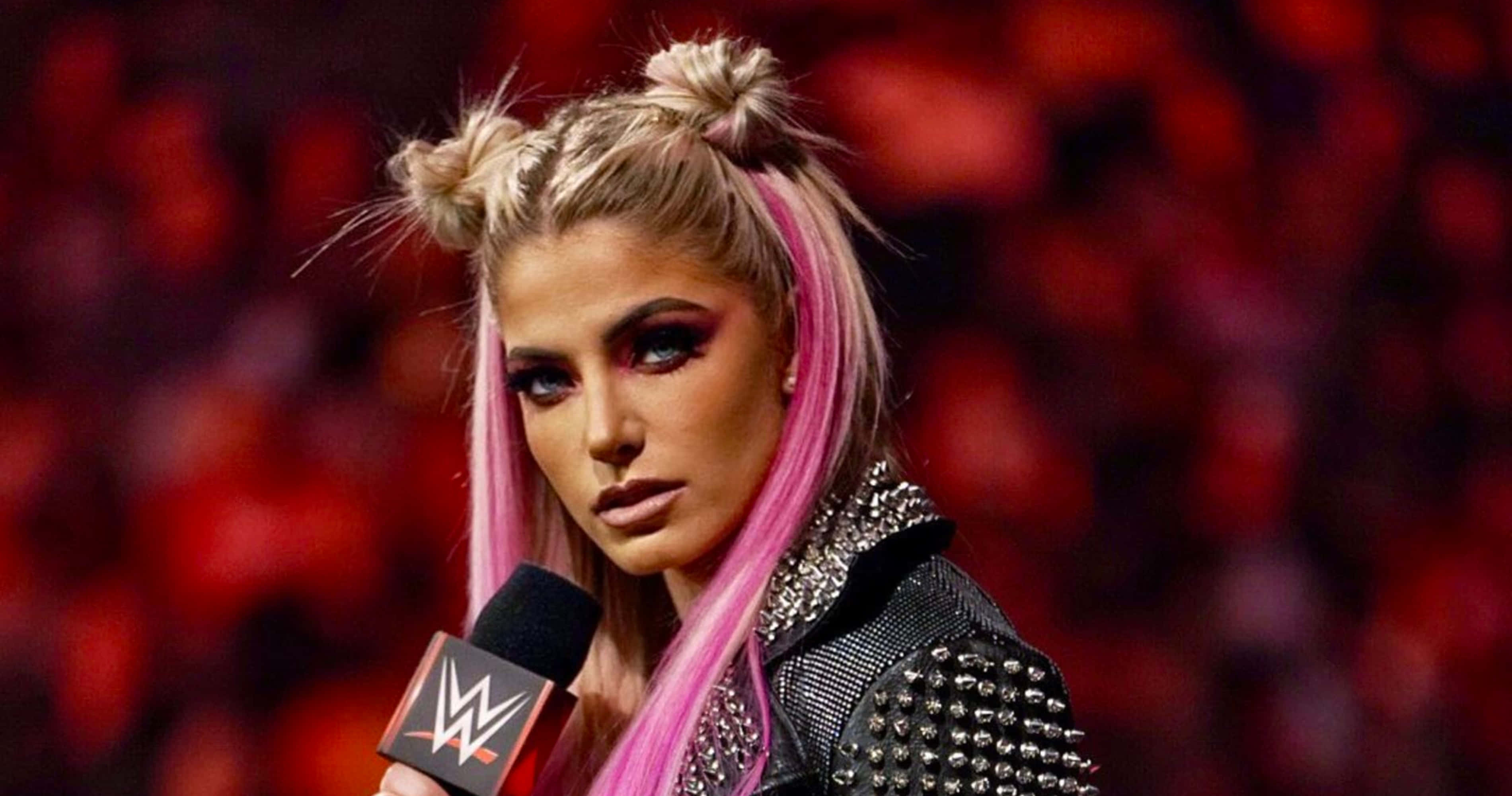 Alexa Bliss 桌布 壁紙 — 一位WWE摔角手，粉紅色和金色頭髮紮成雙丸子頭，在模糊的紅色人群前手持麥克風。