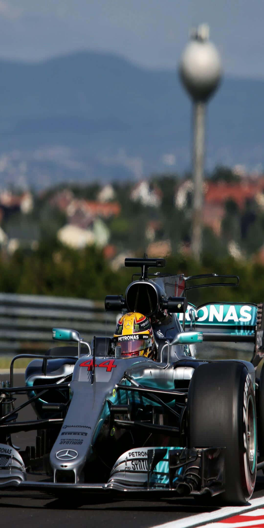 F1 桌布 壁紙 — 一位一級方程式車手駕駛銀色和藍綠色的賓士賽車，在背景有山脈和水塔的賽道上比賽。