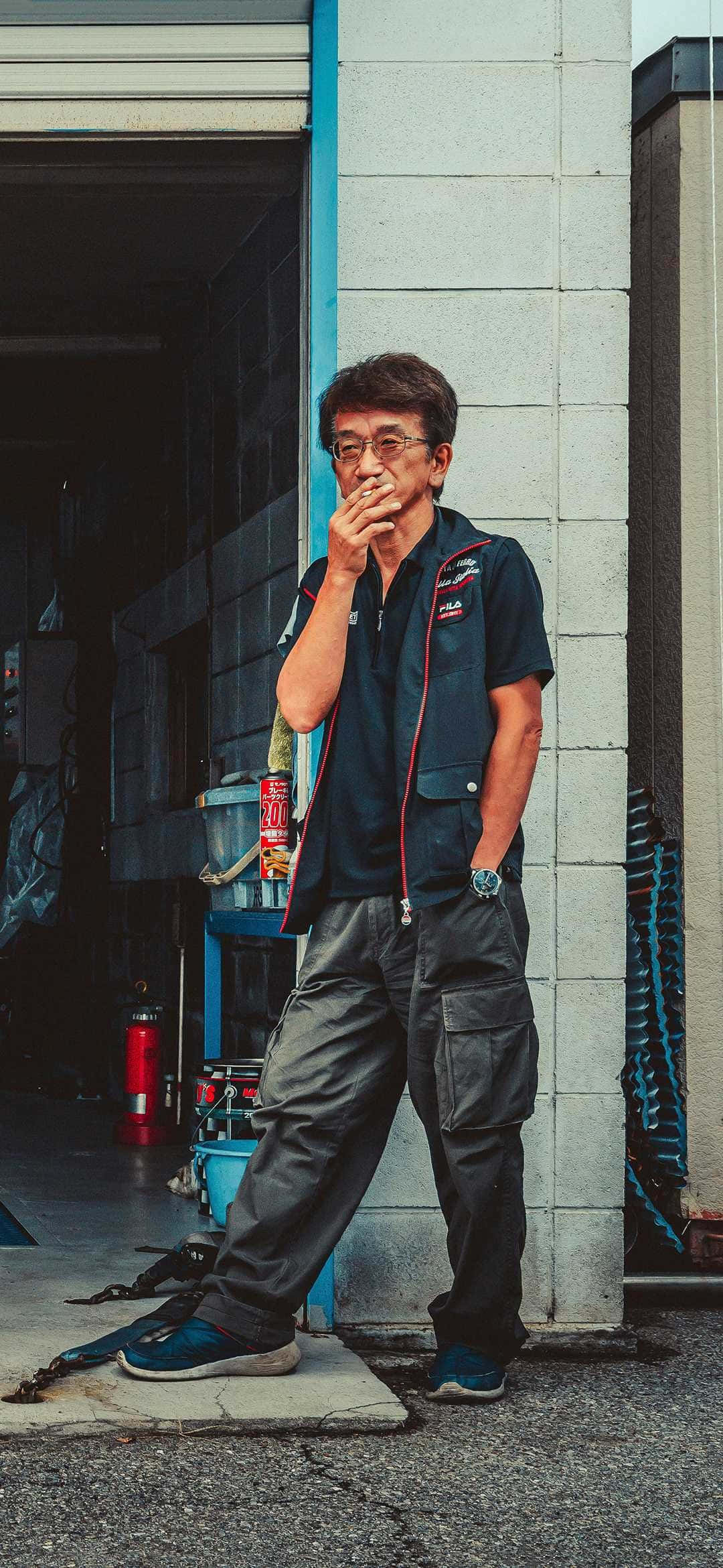 人像動態 桌布 壁紙 — 一位中年機械師穿著深色背心和工裝褲，靠在車庫牆上抽煙。