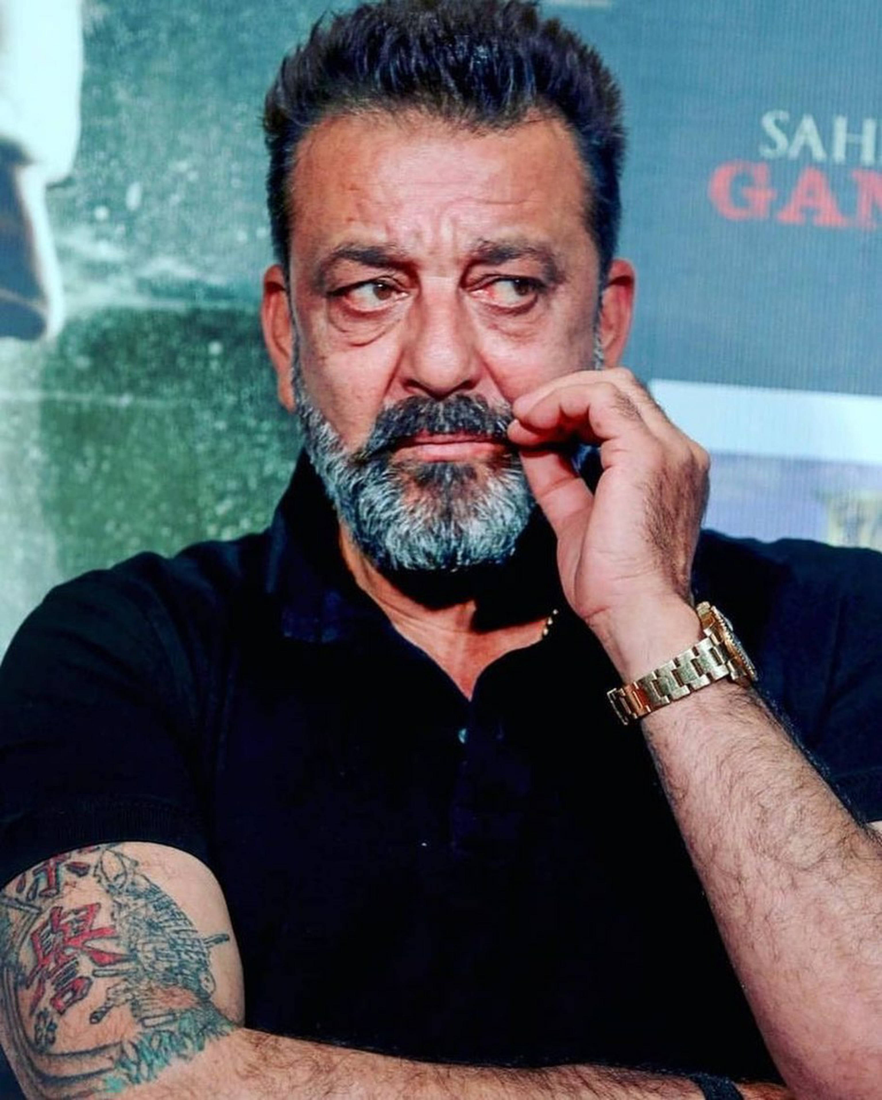 Sanjay 桌布 壁紙 — 一位中年男子，留著灰白鬍鬚，手臂有刺青，在模糊的宣傳背景前用手托著下巴。