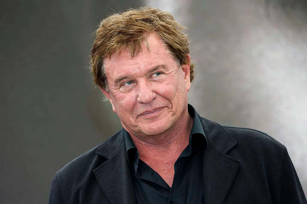 Tom Berenger 桌布 壁紙 — 一位中年男子的特寫肖像，他有亂糟糟的棕色頭髮和微妙的微笑，穿著黑色外套，背景是柔和的灰色。