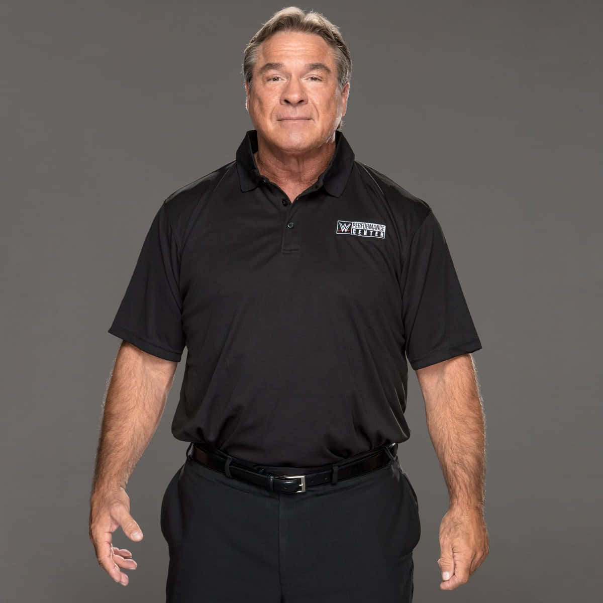 WWE 桌布 壁紙 — 一位中年男子穿著帶有標誌的黑色Polo衫，自信地站在純灰色工作室背景前。