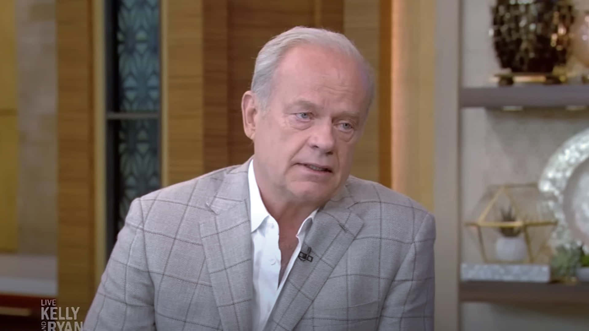 Kelsey Grammer 桌布 壁紙 — 一位中年男子穿著灰色格紋西裝，坐在燈光溫暖的工作室裡，身後有裝飾架。