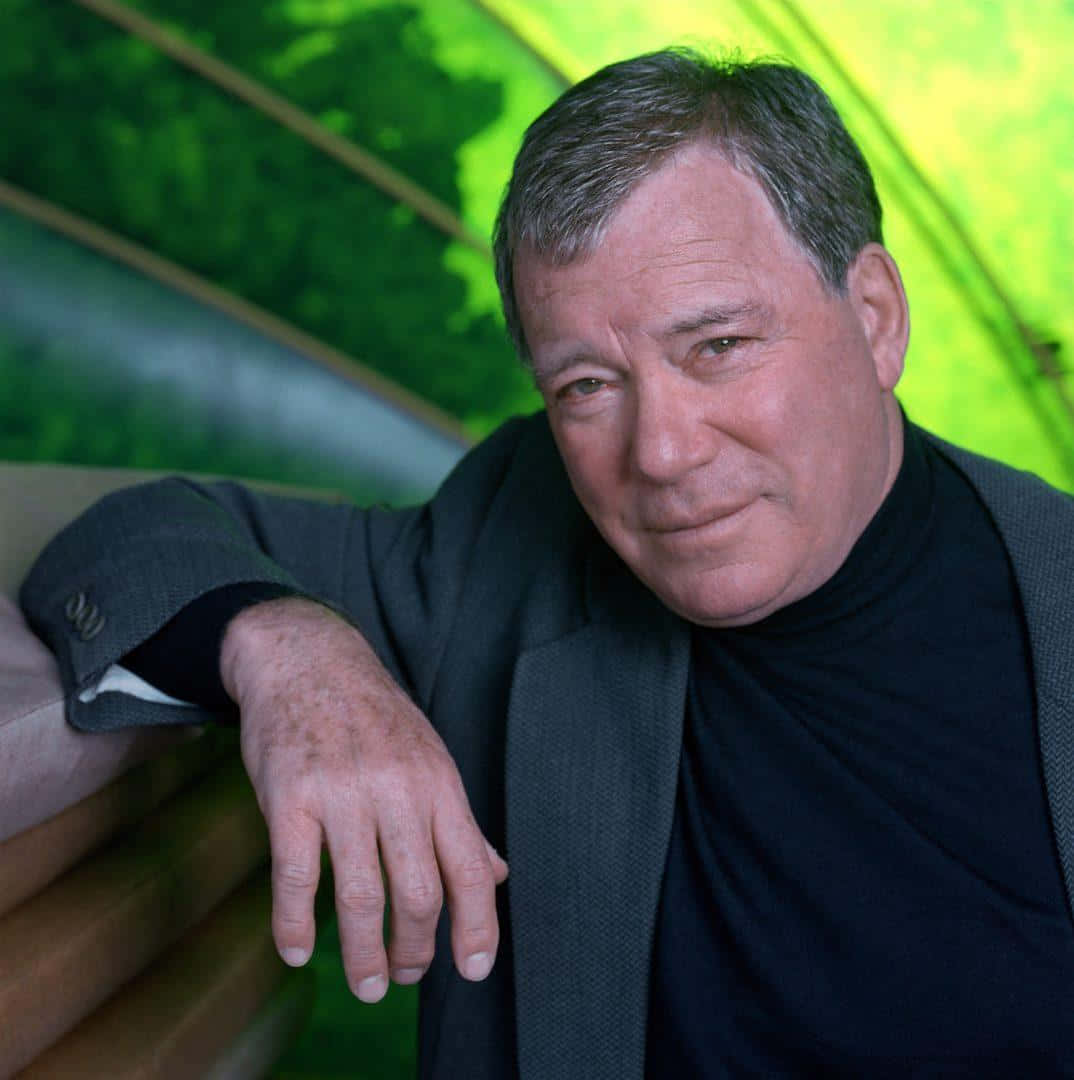 william shatner 桌布 壁紙 — 一位中年男子穿著黑色高領毛衣和西裝外套，將手臂擱在木質表面上，背景是充滿活力的綠色弧形背景。