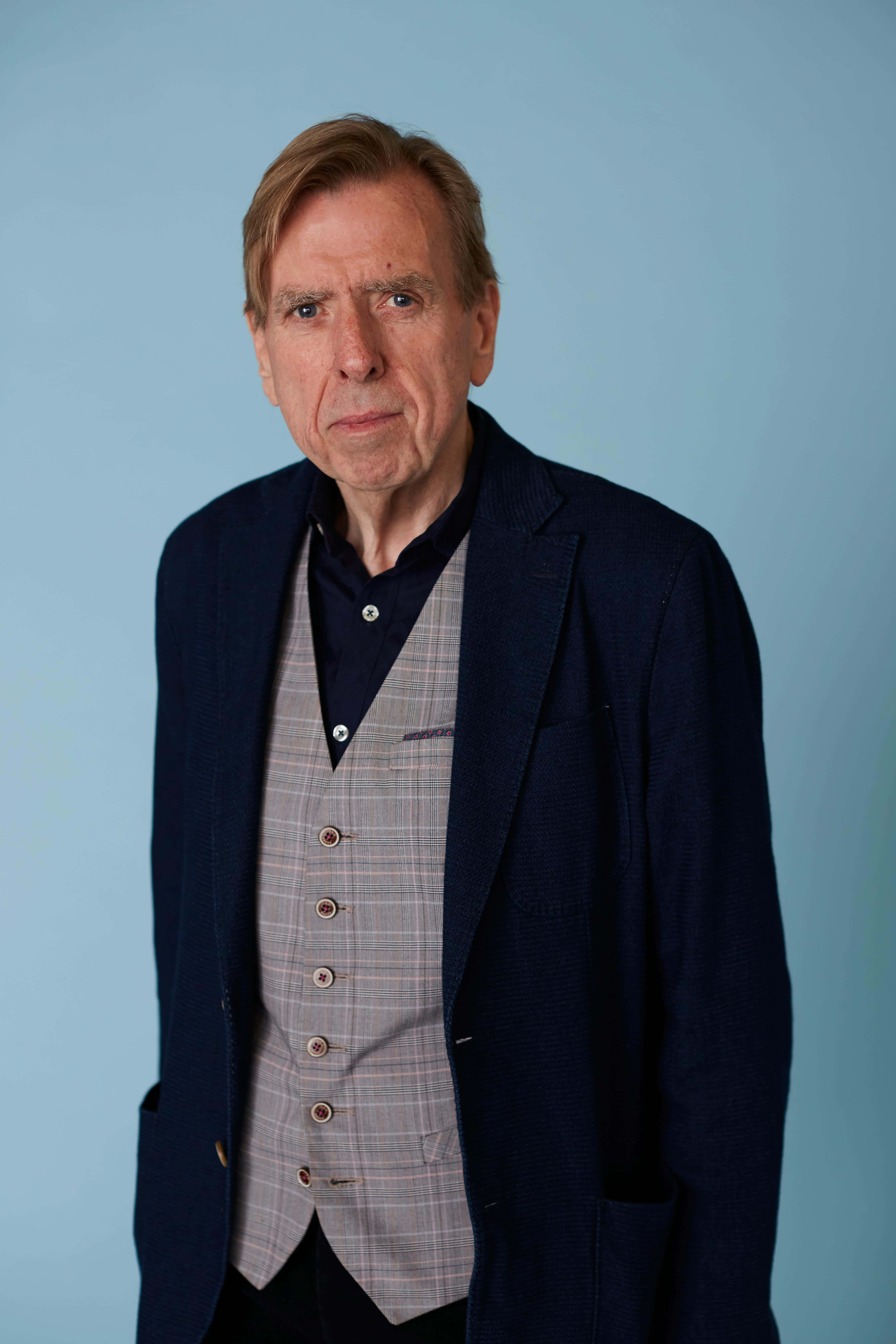 Timothy Spall 桌布 壁紙 — 一位中年男子身穿海軍藍西裝外套和格紋背心，站在純淺藍色攝影棚背景前，表情嚴肅。