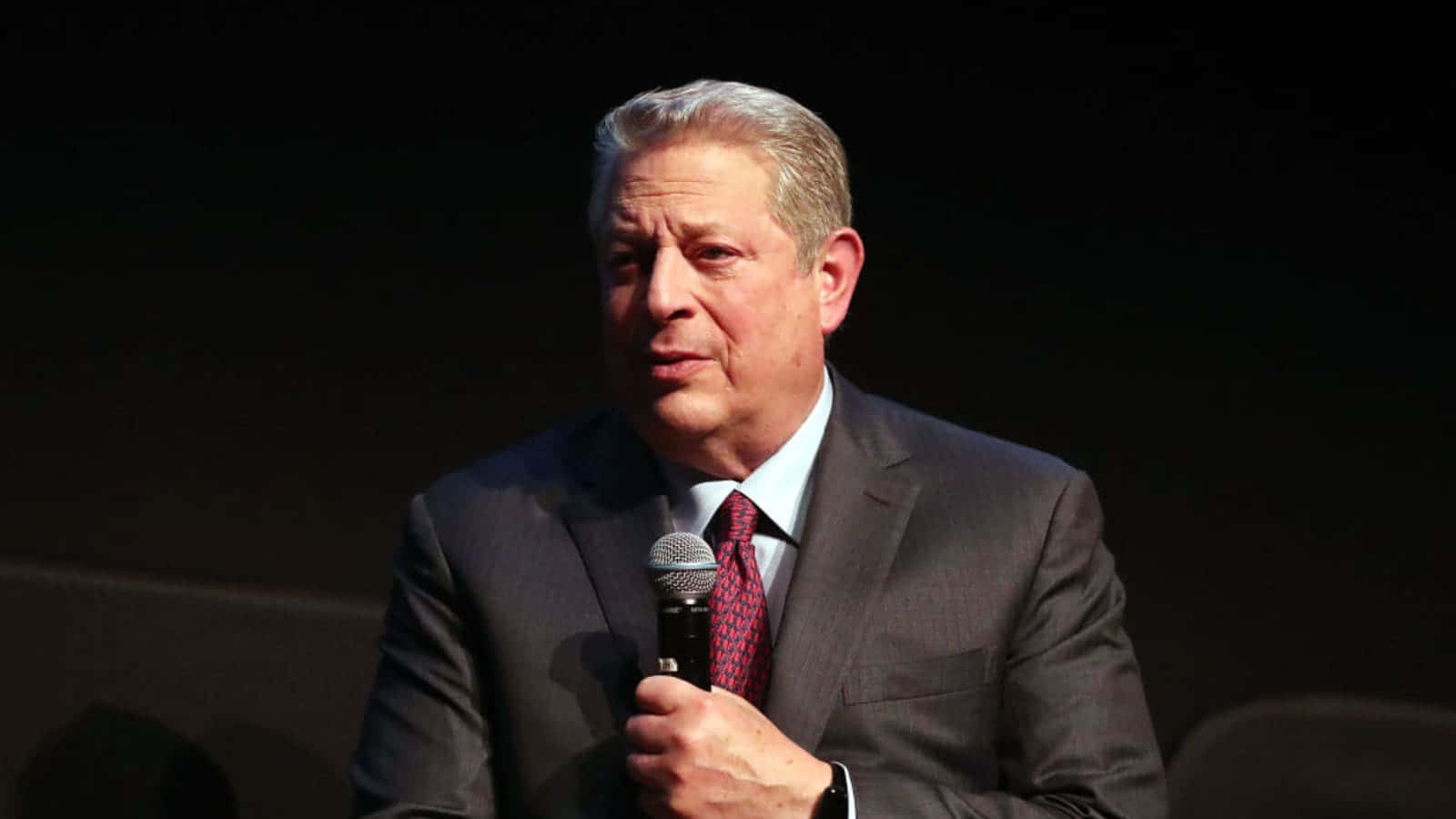Celebrity 桌布 壁紙 — 一位中年男子身穿深色西裝、打紅色領帶，在黑色背景前對著麥克風說話。