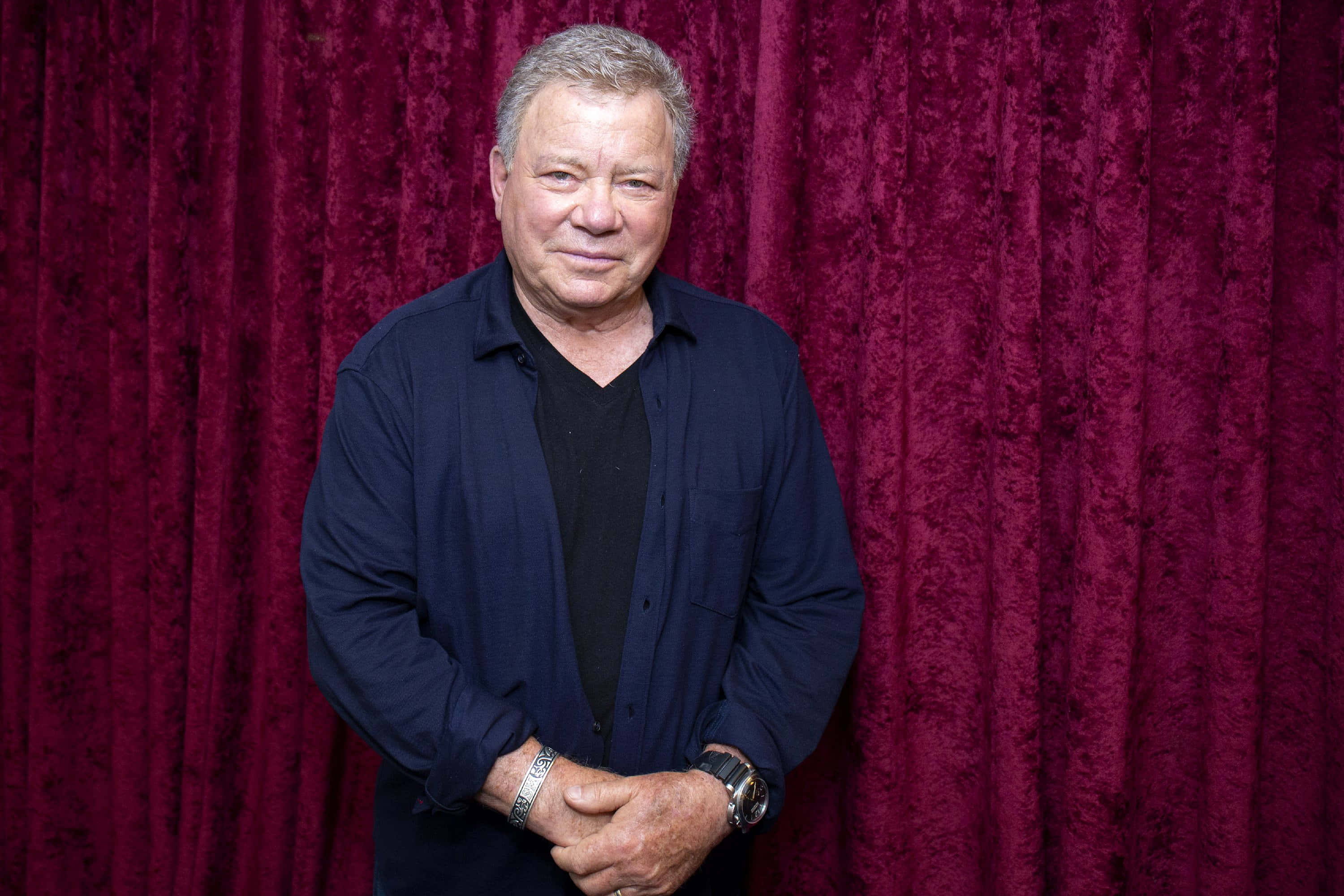 william shatner 桌布 壁紙 — 一位中年男子身穿深藍色襯衫，自信地站在華麗的勃艮第酒紅色天鵝絨窗簾前。