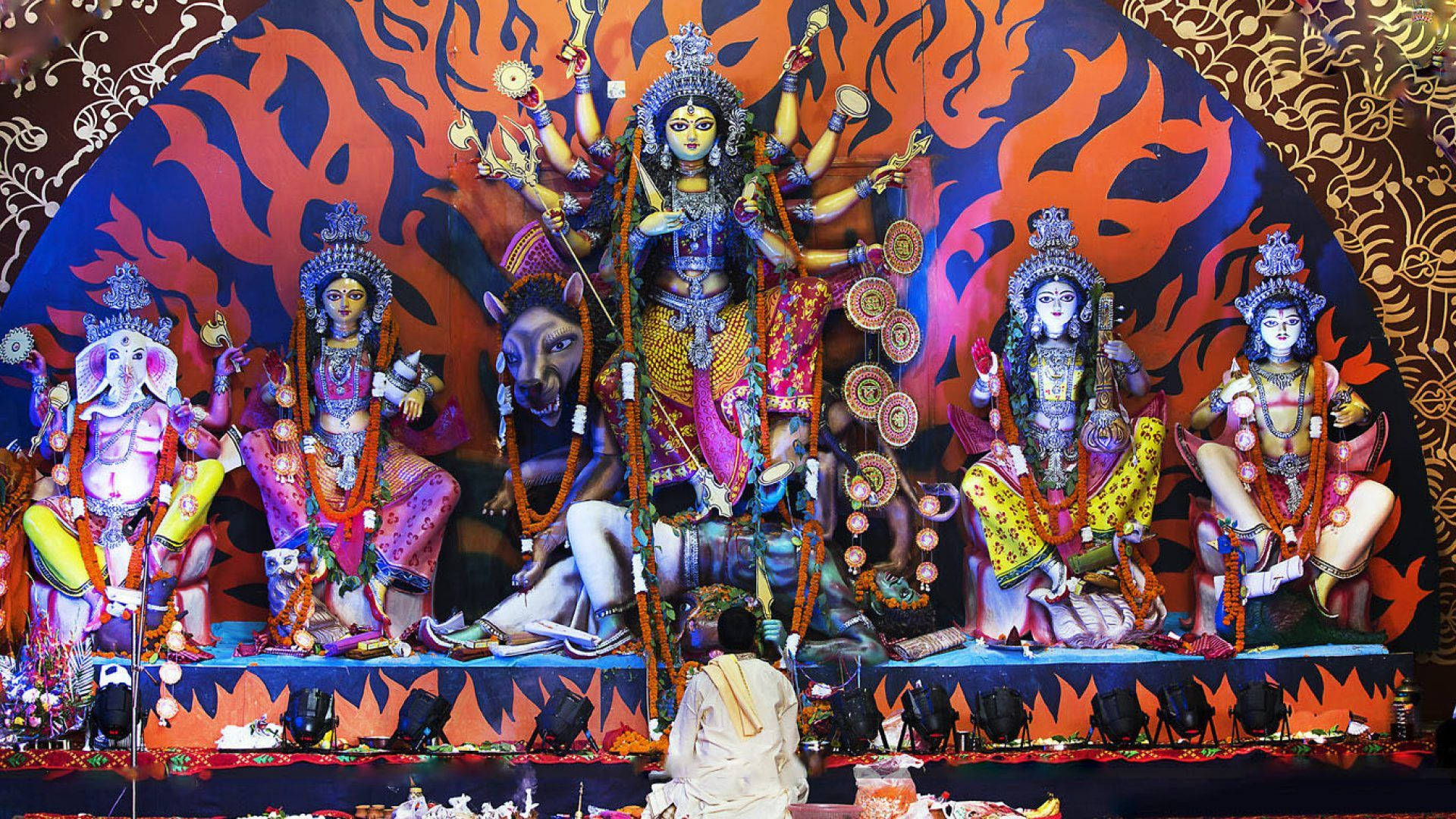 durga mata HD 桌布 壁紙 — 一位信徒在熾熱的橙色和藍色背景下，向宏偉多彩的杜爾迦神像鞠躬，神像周圍環繞著她的神聖侍從。