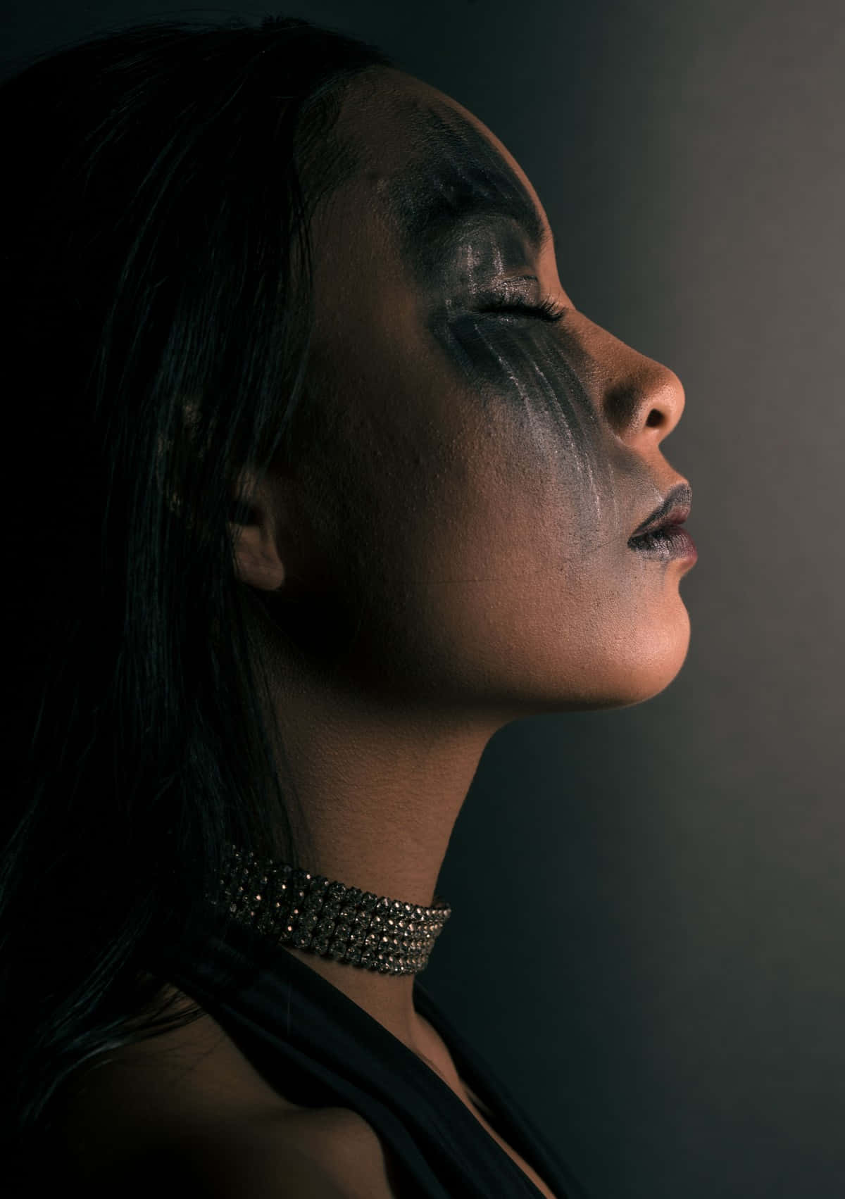 Women Profile 桌布 壁紙 — 一位側面輪廓的女性，臉上帶有戲劇性的黑色和銀色顏料，戴著閃閃發光的頸鏈，背景為深色。