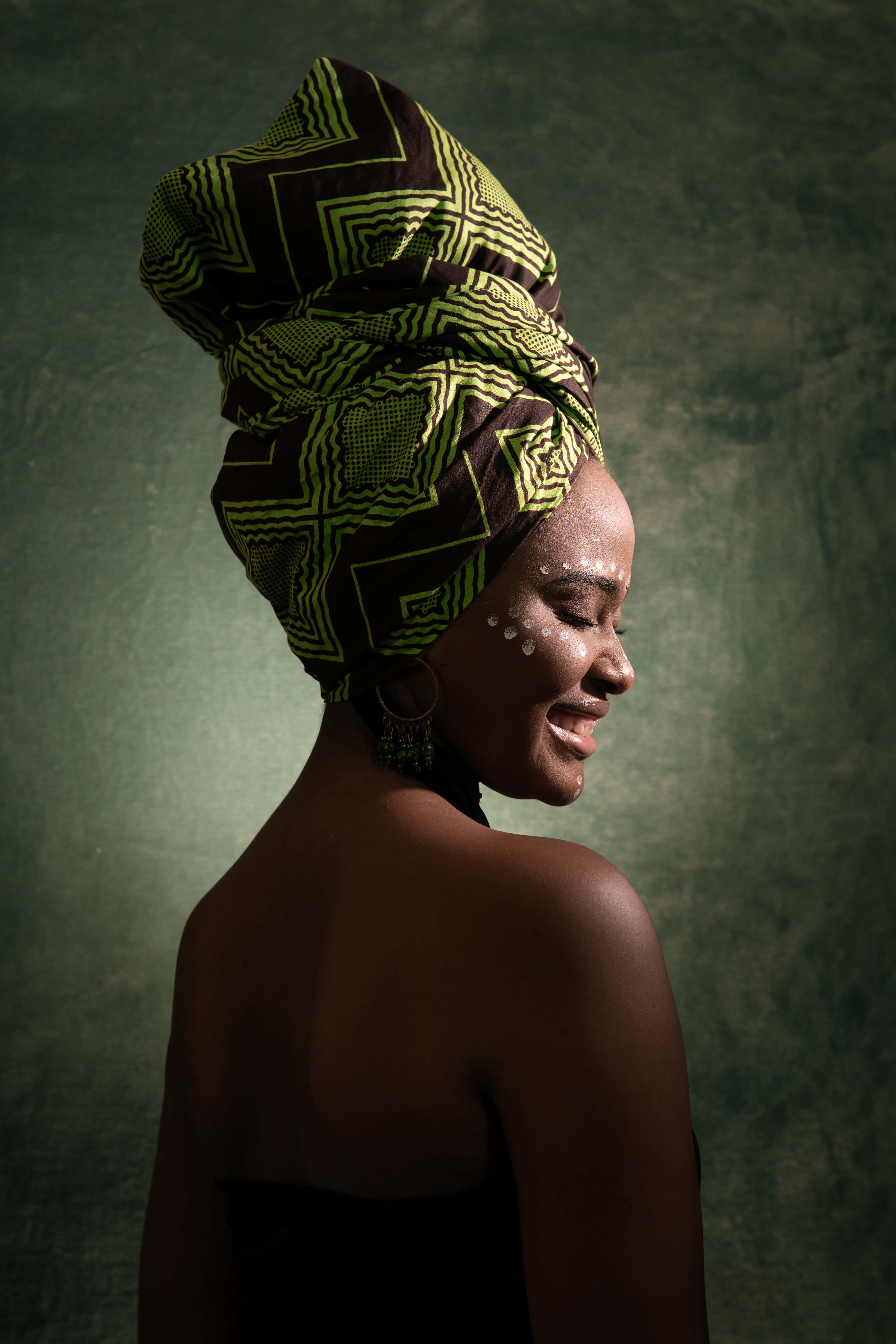 African 桌布 壁紙 — 一位側面輪廓的女性戴著充滿活力的綠色和棕色圖案頭巾，在紋理綠色背景前溫柔微笑。