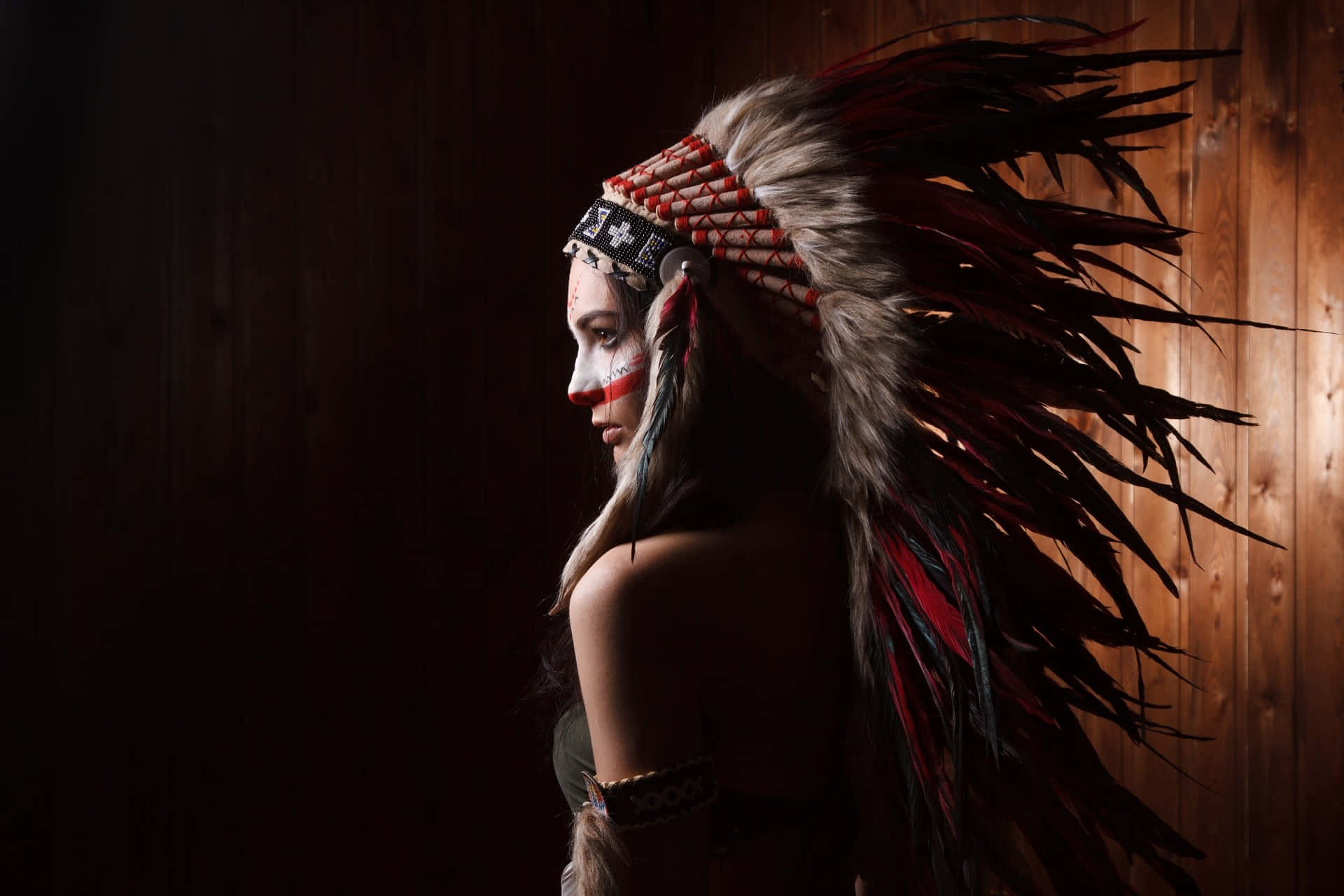 Tribal 桌布 壁紙 — 一位側面輪廓的女性戴著戲劇性的羽毛頭飾和臉部彩繪，背景是深色木牆。