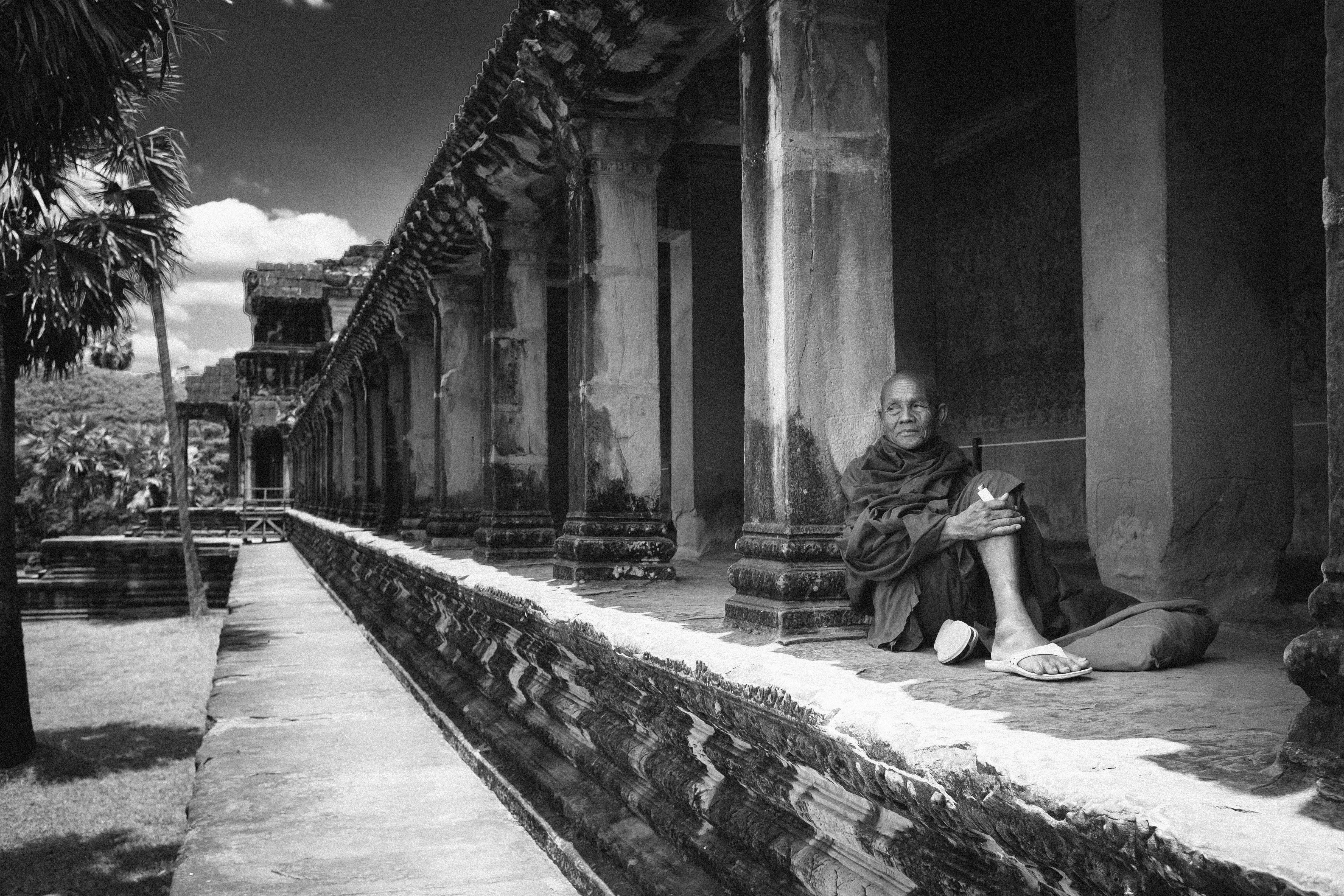 Cambodia 桌布 壁紙 — 一位僧侶平靜地坐在風化的石台上，下方是古老的寺廟柱子，場景為單色調。
