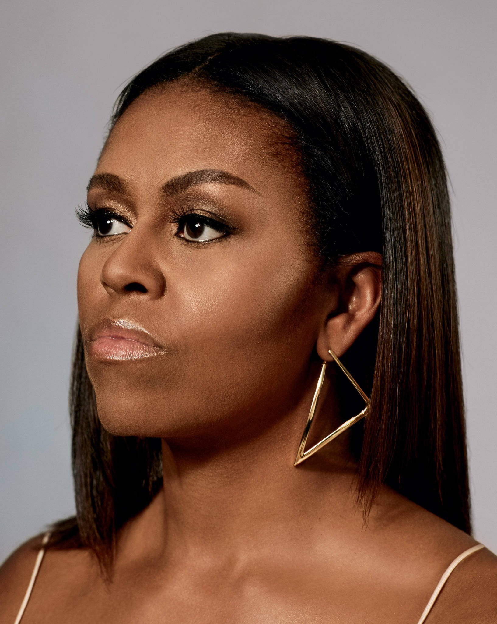 Michelle 桌布 壁紙 — 一位優雅的女人，擁有光滑的黑髮和大膽的幾何金色耳環，在柔和的灰色背景下若有所思地凝視。