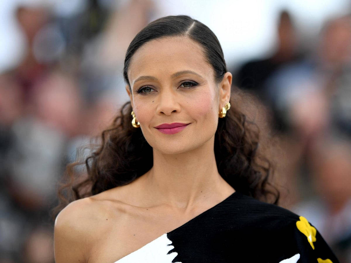 Actress HD 桌布 壁紙 — 一位優雅的女人，深色捲髮，戴著金色耳環，在擁擠的活動中穿著黑白單肩裙，溫柔地微笑。