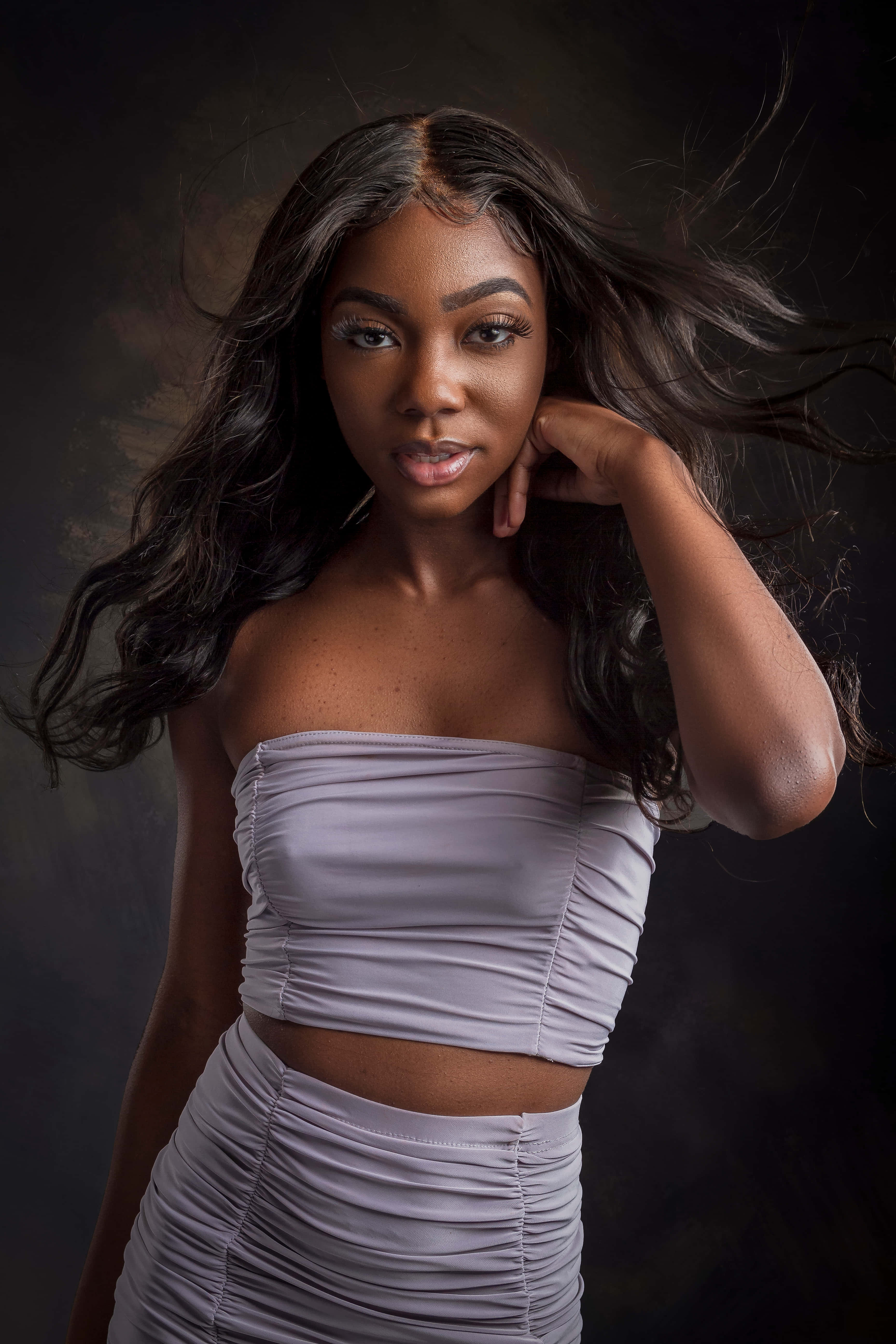Beautiful Black Woman 桌布 壁紙 — 一位優雅的女人，飄逸的深色頭髮，穿著皺褶薰衣草色兩件套，背景陰暗。