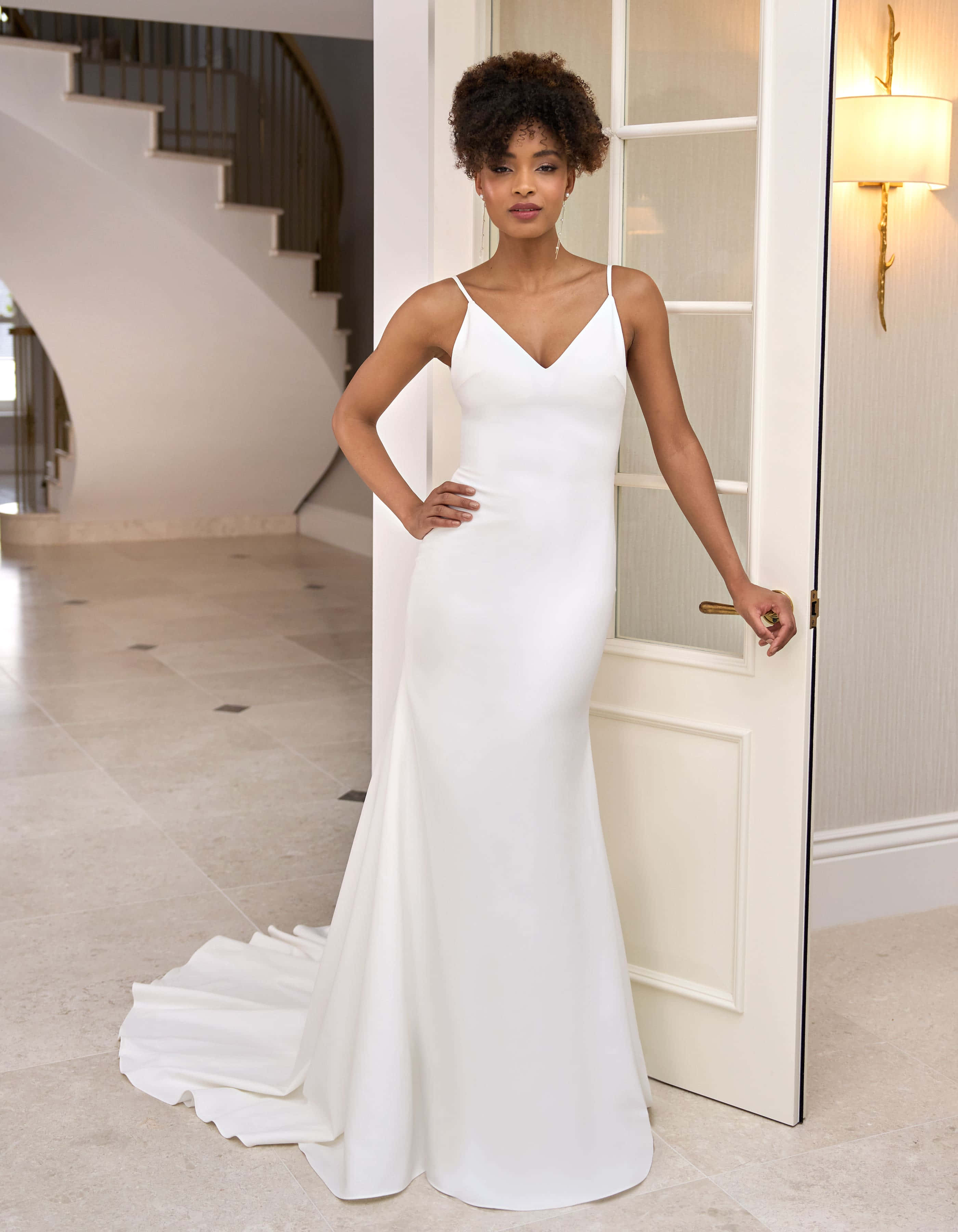 Bride's Dress 桌布 壁紙 — 一位優雅的女人穿著一件修身的白色婚紗，在一個有弧形樓梯的現代住宅中，優雅地站在一扇打開的門旁邊。