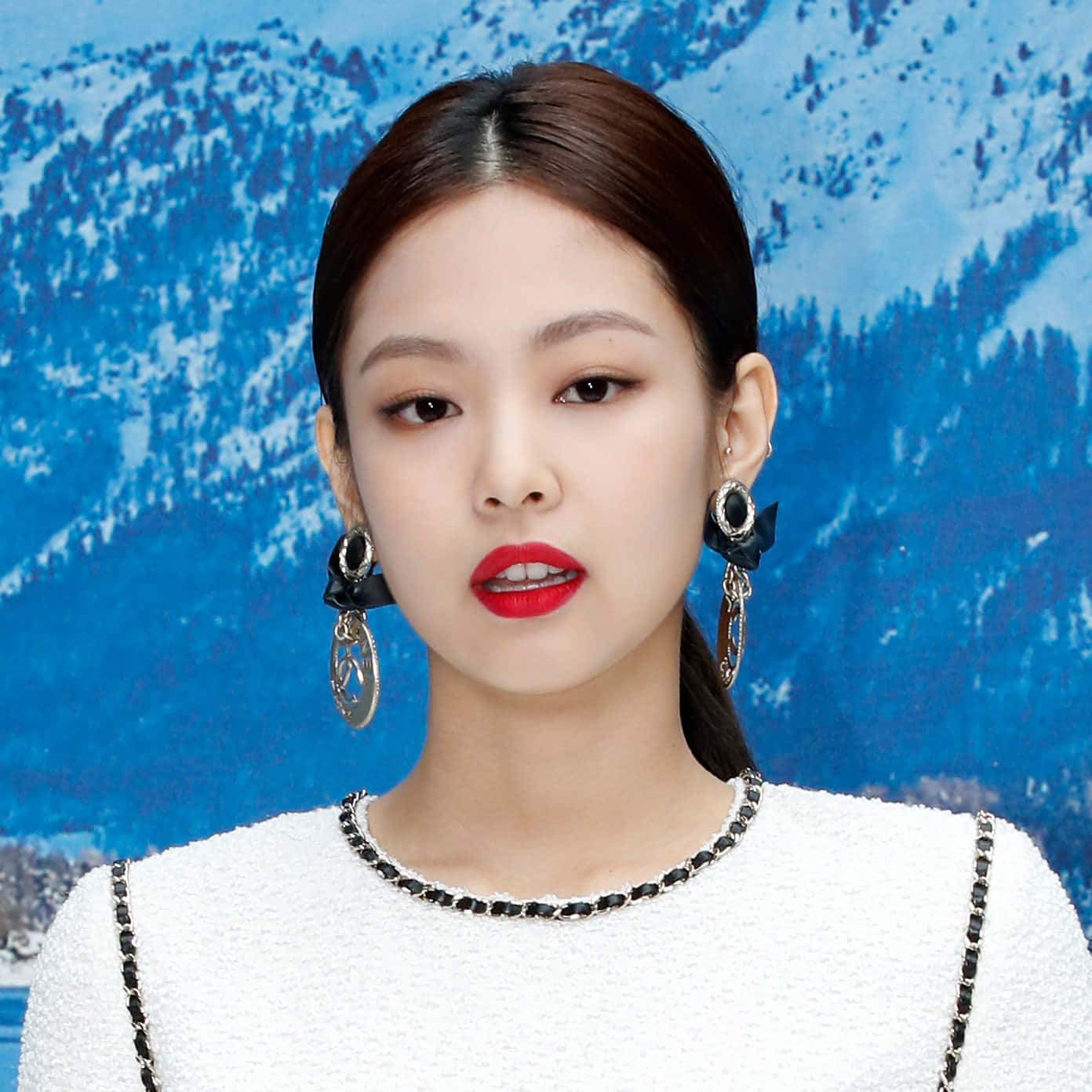 Jennie 桌布 壁紙 — 一位優雅的女士，塗上大膽的紅唇，戴著搶眼的耳環，站在白雪覆蓋的山景前面。
