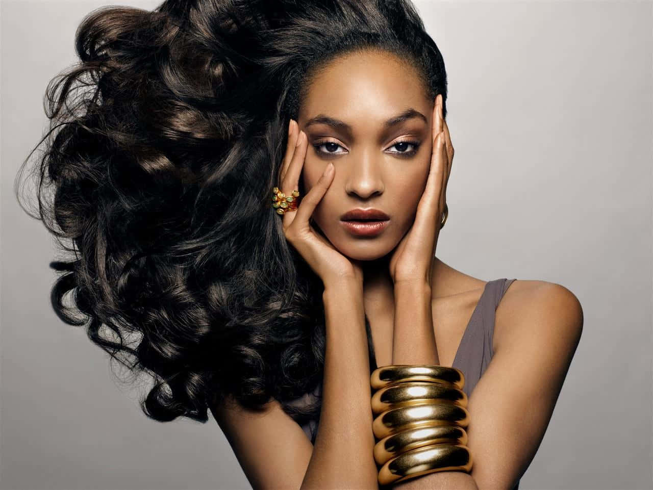 jourdan dunn 桌布 壁紙 — 一位優雅的女士，濃密的深色捲髮勾勒出她的臉龐，她將雙手放在飾有金飾的臉頰上。