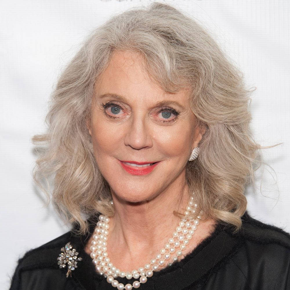 Celebrity 桌布 壁紙 — 一位優雅的女士，銀灰色波浪長髮，身穿黑色服裝，飾有珍珠和閃閃發光的胸針，背景為白色。