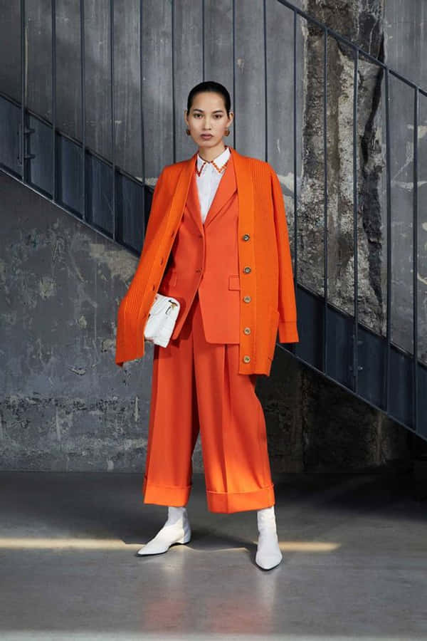 Clothes 桌布 壁紙 — 一位優雅的女士穿著大膽的橙色西裝，自信地站在一個有金屬欄杆的風化工業牆前面。