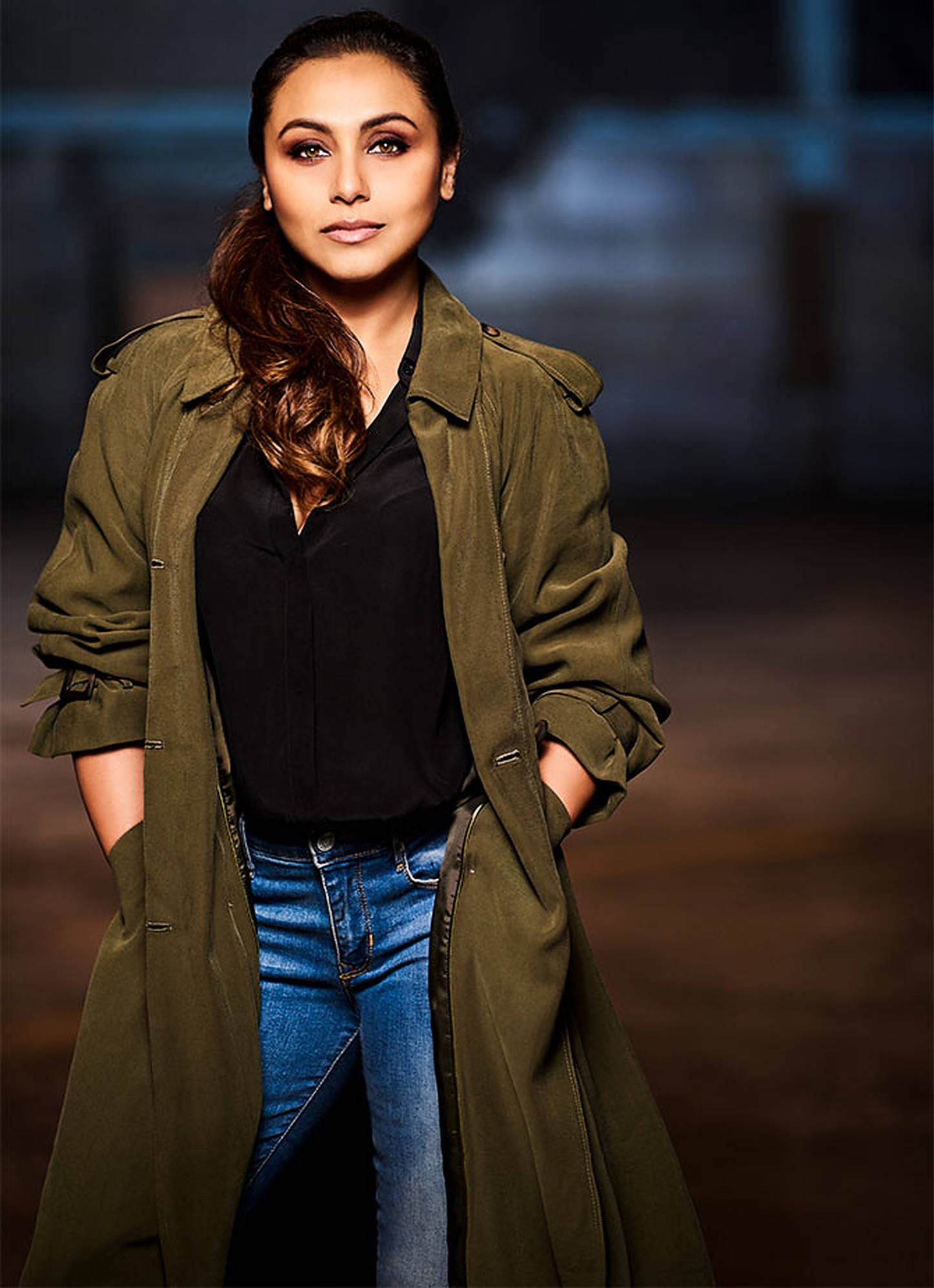 Rani Mukerji 桌布 壁紙 — 一位優雅的女士穿著橄欖色風衣和牛仔褲，雙手插袋，自信地站在模糊的城市背景前。