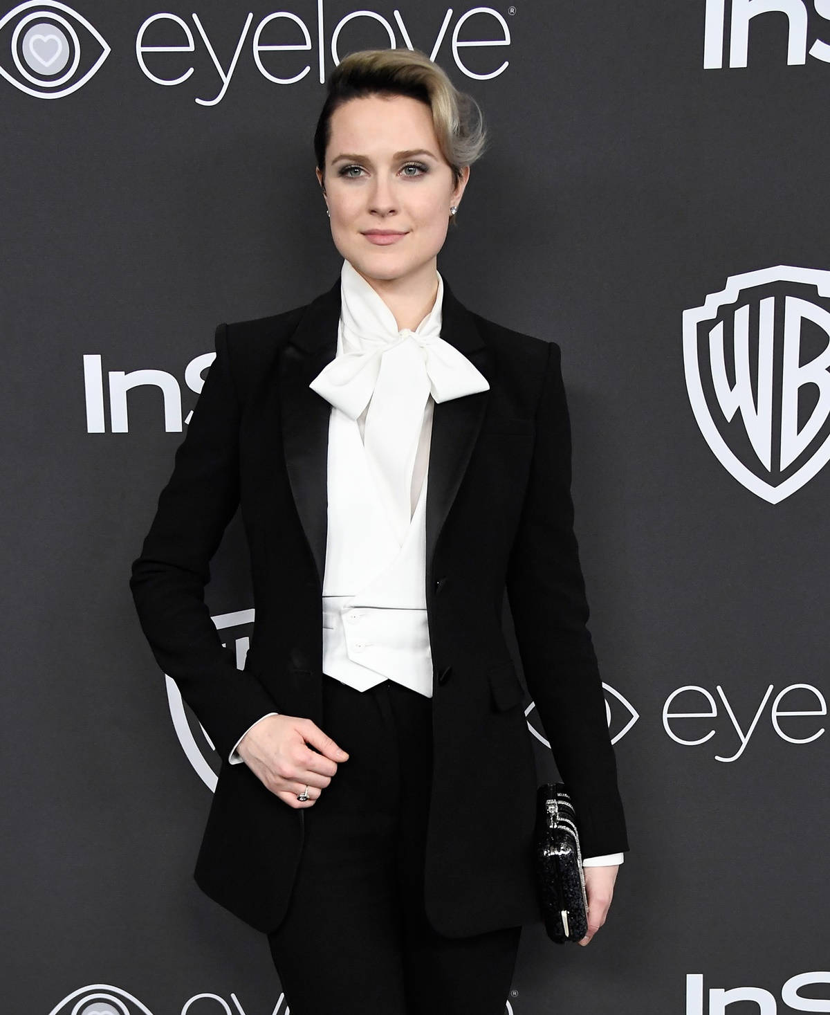 Celebrity 桌布 壁紙 — 一位優雅的女士身穿俐落黑色燕尾服配白色領結，自信地站在品牌活動背景前。