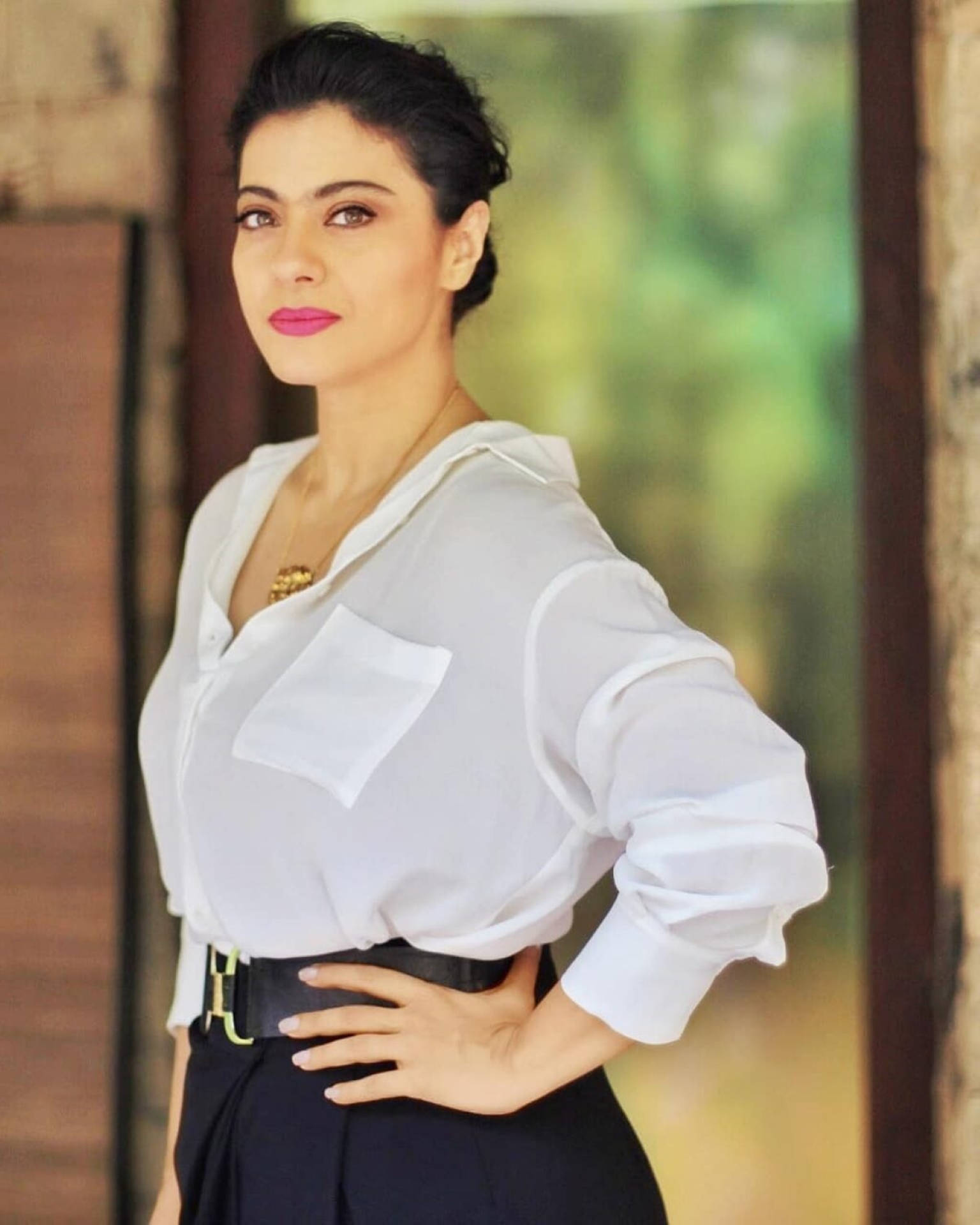 Kajol 桌布 壁紙 — 一位優雅的女士身穿白色襯衫和黑色裙子，一手叉腰，自信地站在模糊的綠色背景前。