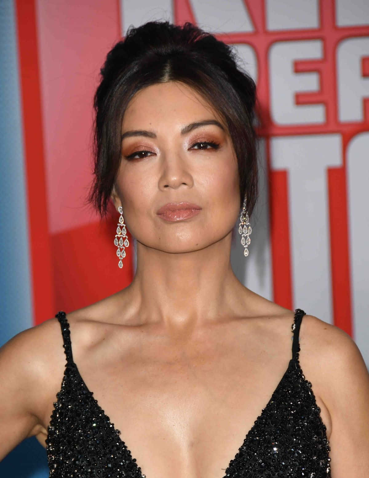 Ming Na Wen 桌布 壁紙 — 一位優雅的女士身穿閃亮黑色連衣裙，戴著垂墜耳環，在紅白活動背景前擺姿勢。