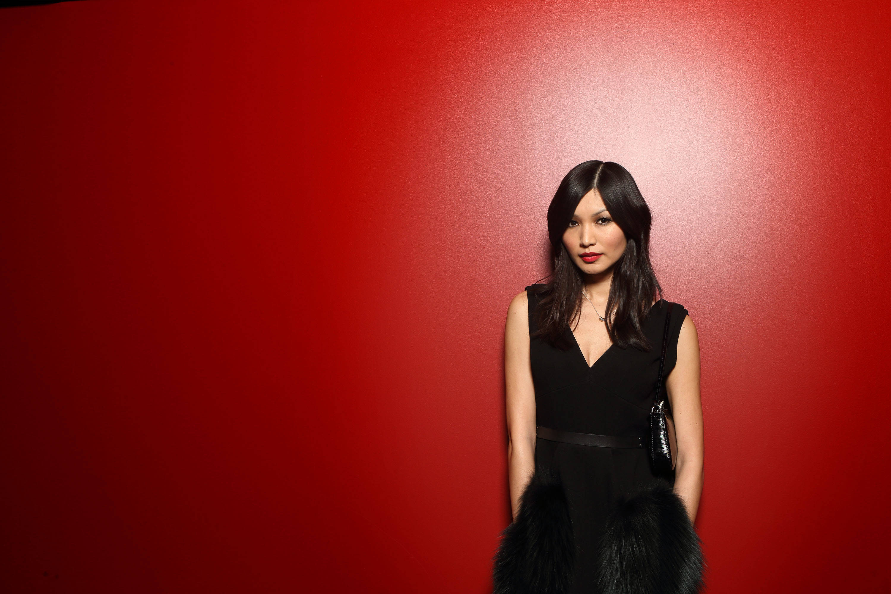 Gemma Chan 桌布 壁紙 — 一位優雅的女士身穿黑色毛邊連衣裙，站在鮮紅色的牆前，散發自信與優雅。