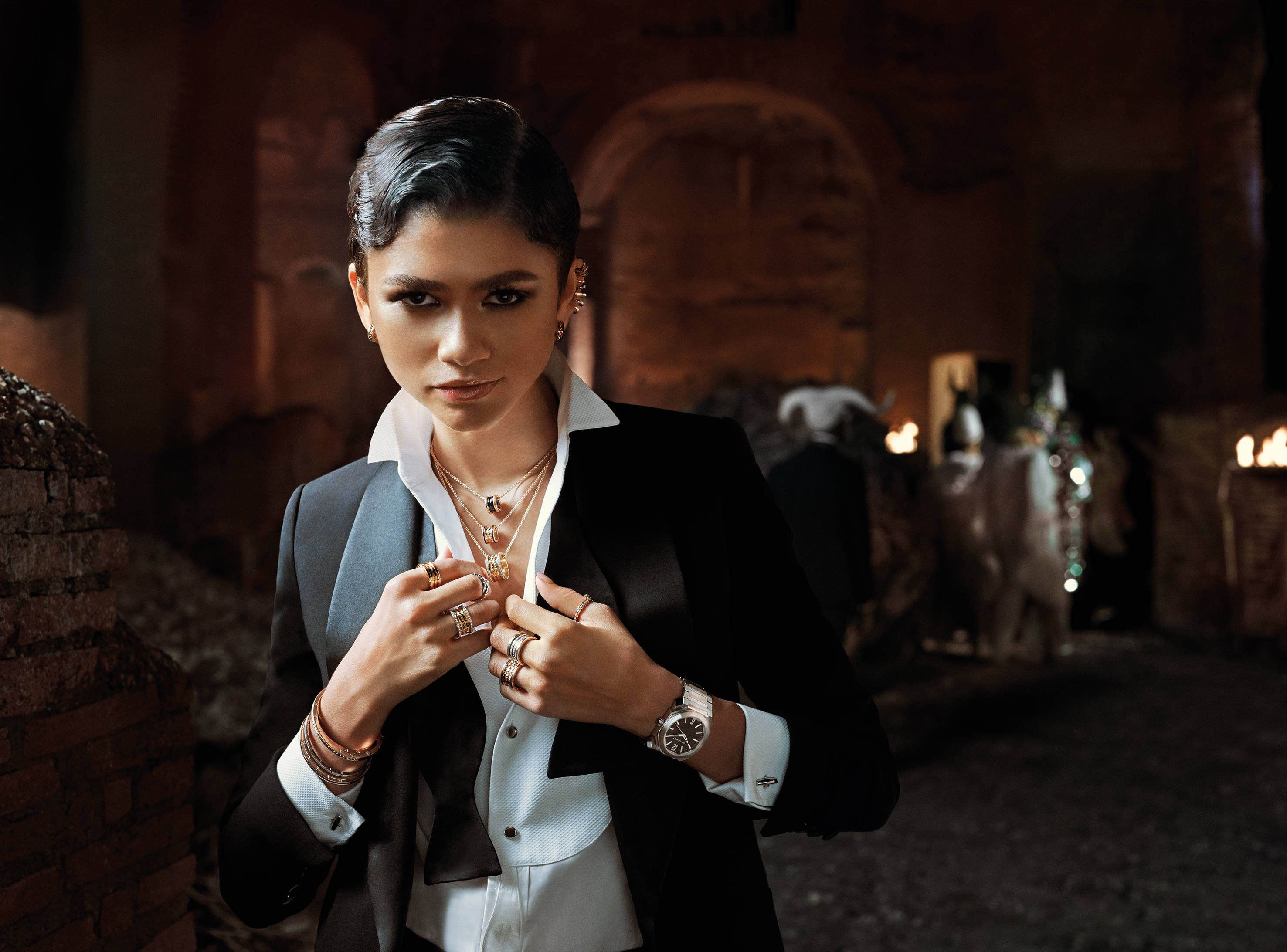 Zendaya 桌布 壁紙 — 一位優雅的女士身穿黑色西裝外套和白色襯衫，在昏暗的古老石拱門前調整她的珠寶。