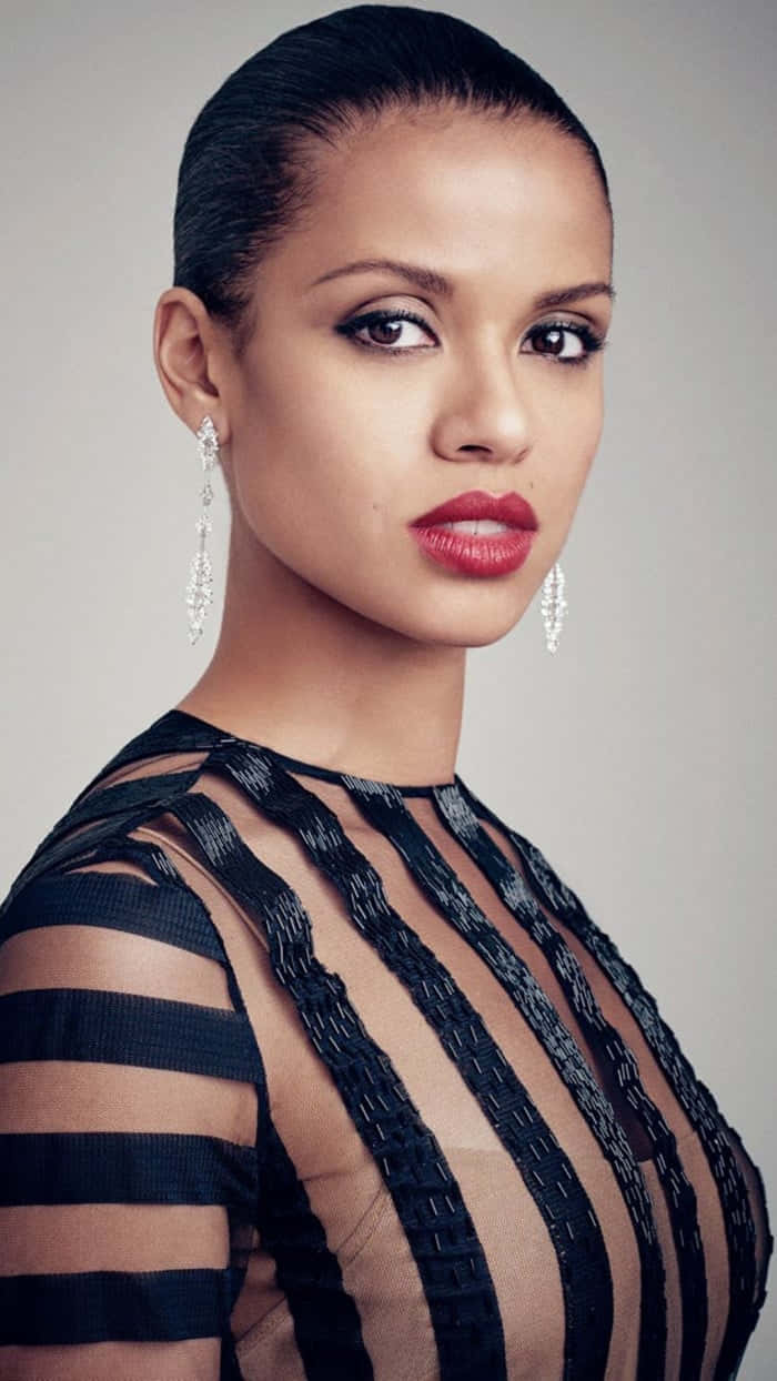 Gugu Mbatha-raw 桌布 壁紙 — 一位優雅的女性，擁有光滑的頭髮和鮮豔的紅唇，穿著黑色條紋透視上衣和閃閃發光的吊燈耳環，背景為中性色調。