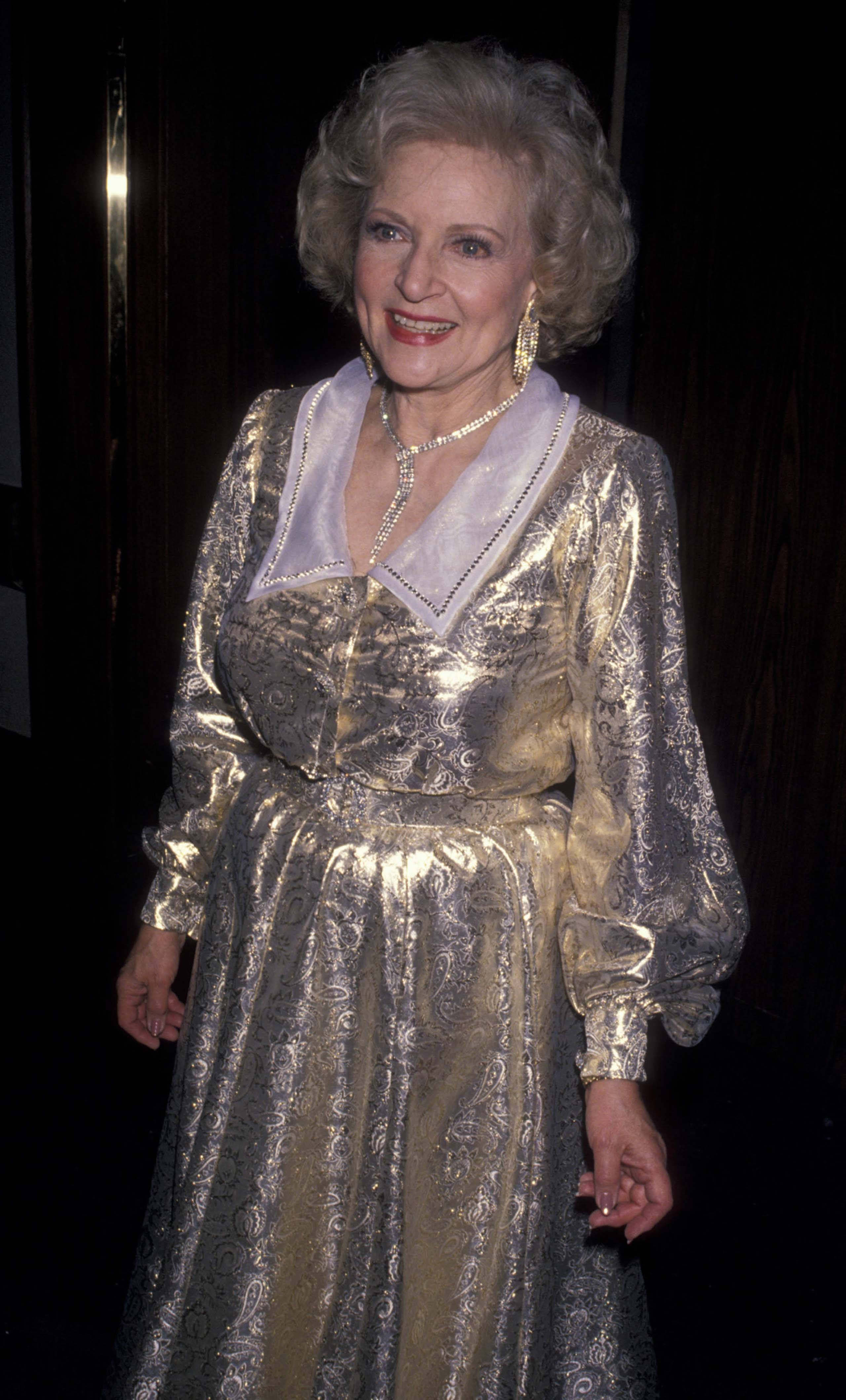 Betty White 桌布 壁紙 — 一位優雅的年長女性穿著閃閃發光的金色和銀色圖案長裙，在黑暗背景下溫暖地微笑。