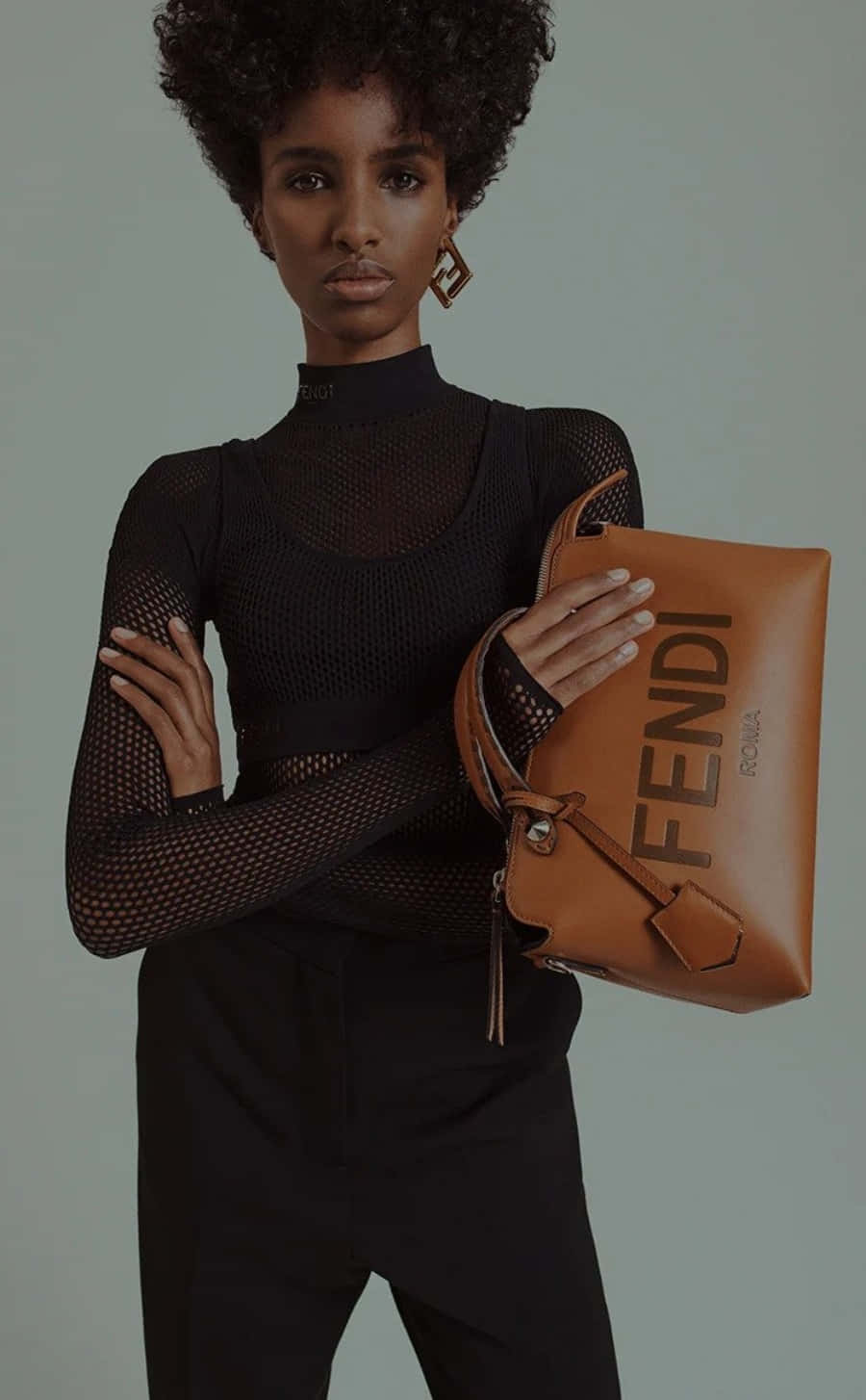 Fendi 桌布 壁紙 — 一位優雅的捲髮女性穿著黑色網狀上衣，在柔和的灰色背景前拿著一個棕褐色的Fendi手袋。