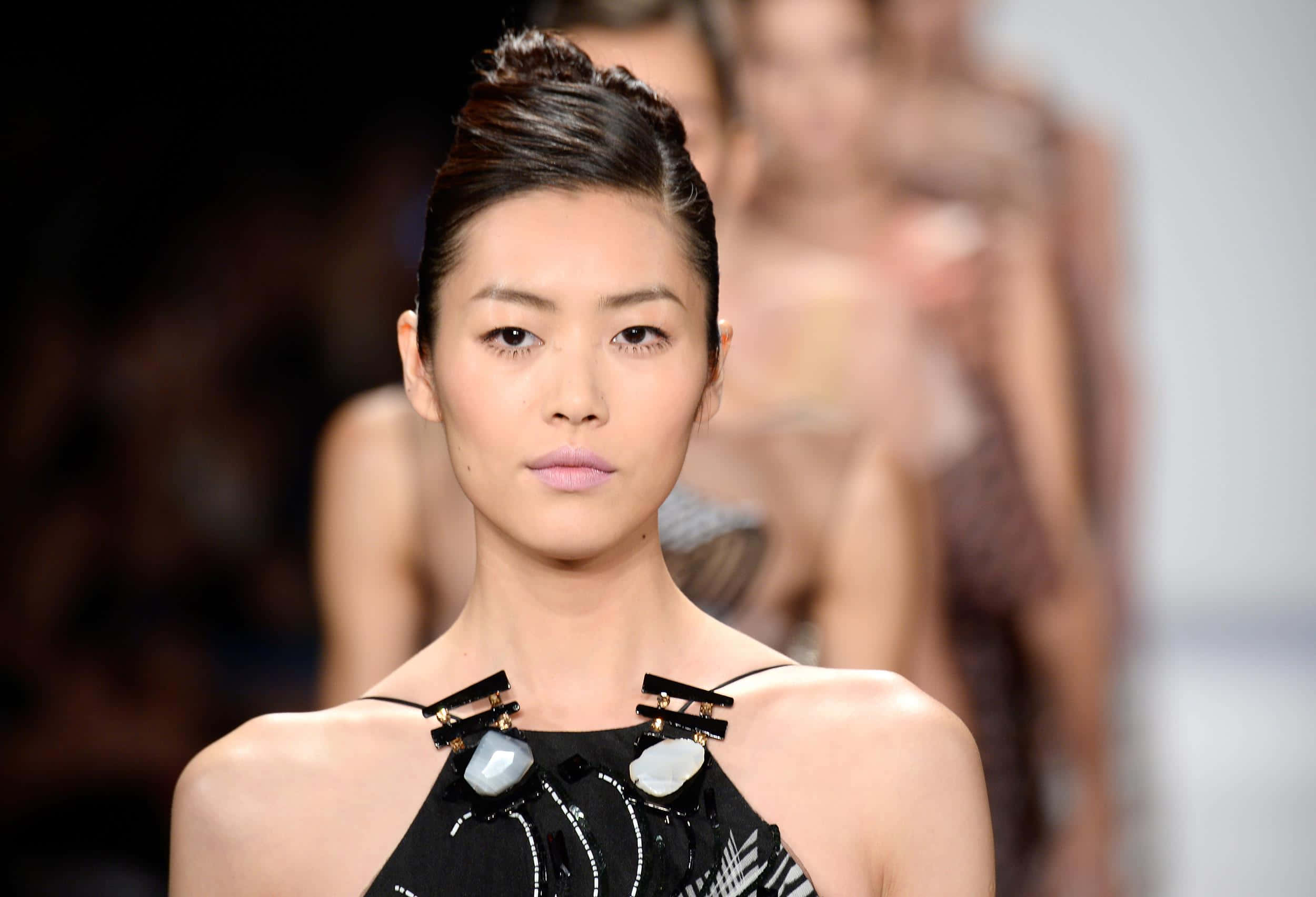 Beautiful Fashion 桌布 壁紙 — 一位優雅的模特兒梳著光滑的髮髻，佩戴大膽的珠寶，在明亮的燈光下自信地走在時裝伸展台上。