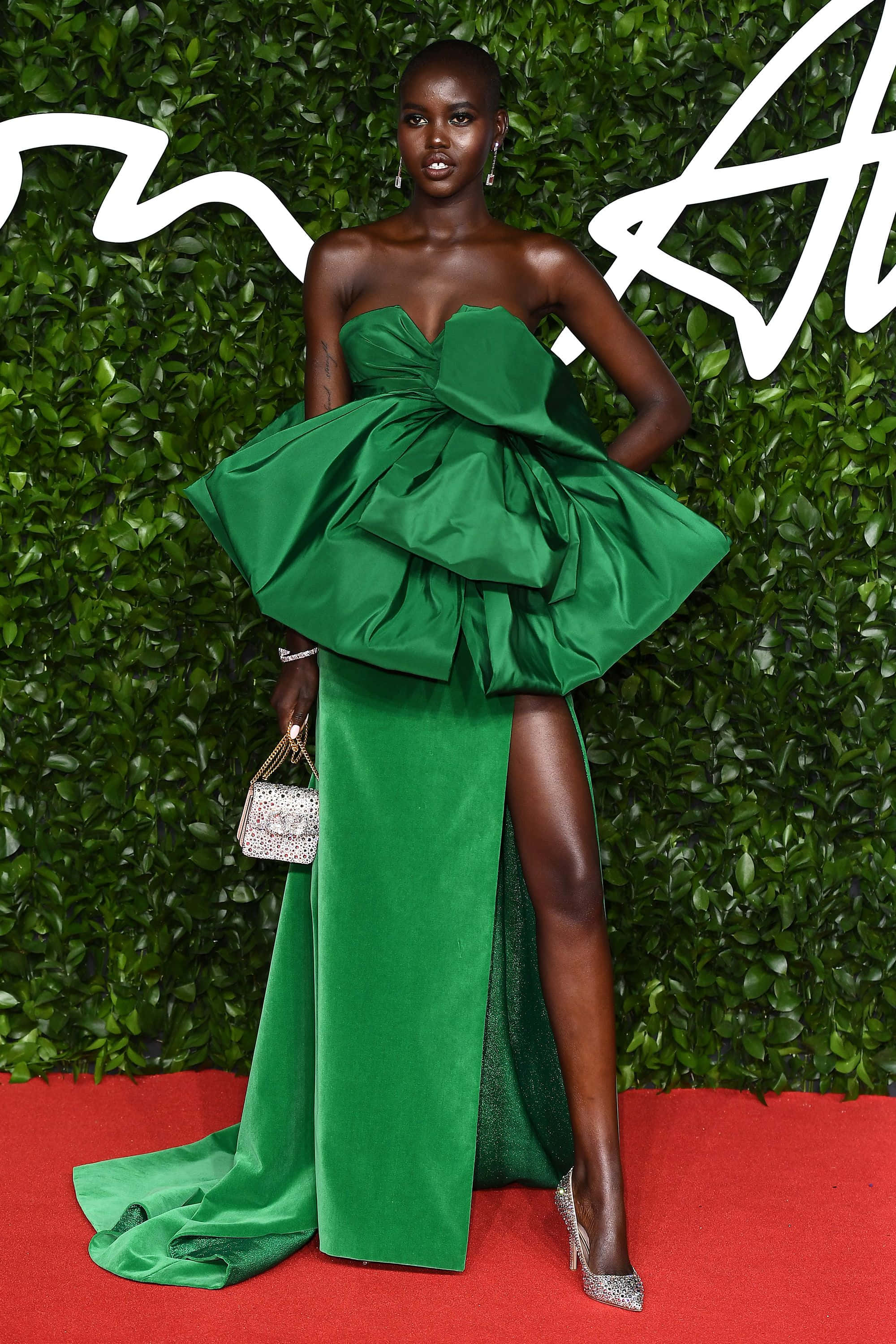 Green Fashion 桌布 壁紙 — 一位優雅的模特兒穿著寬鬆的翡翠綠長裙，自信地站在紅地毯上，背景是茂密的綠色樹籬。