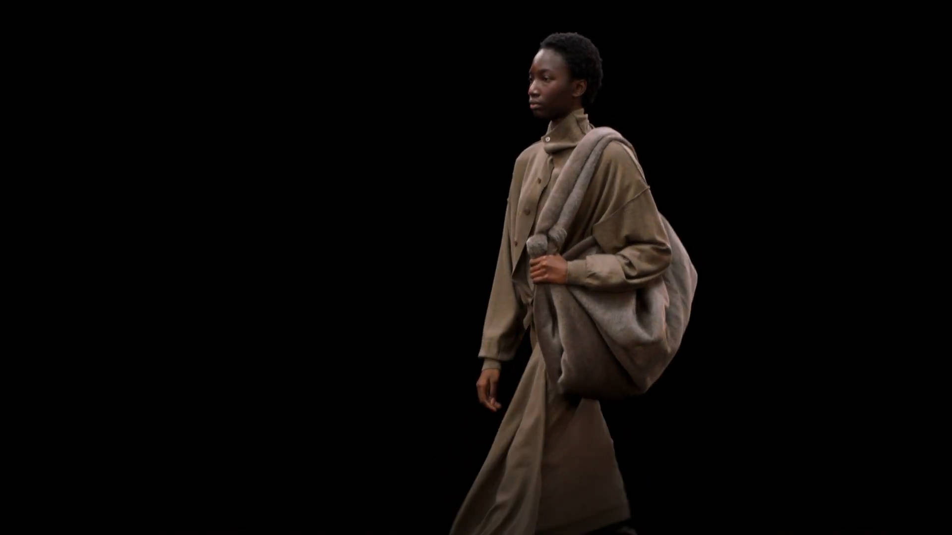 Mens Fashion 桌布 壁紙 — 一位優雅的模特兒穿著飄逸的米色外套，在戲劇性的攝影棚燈光下，自信地走在鮮明的黑色背景前。