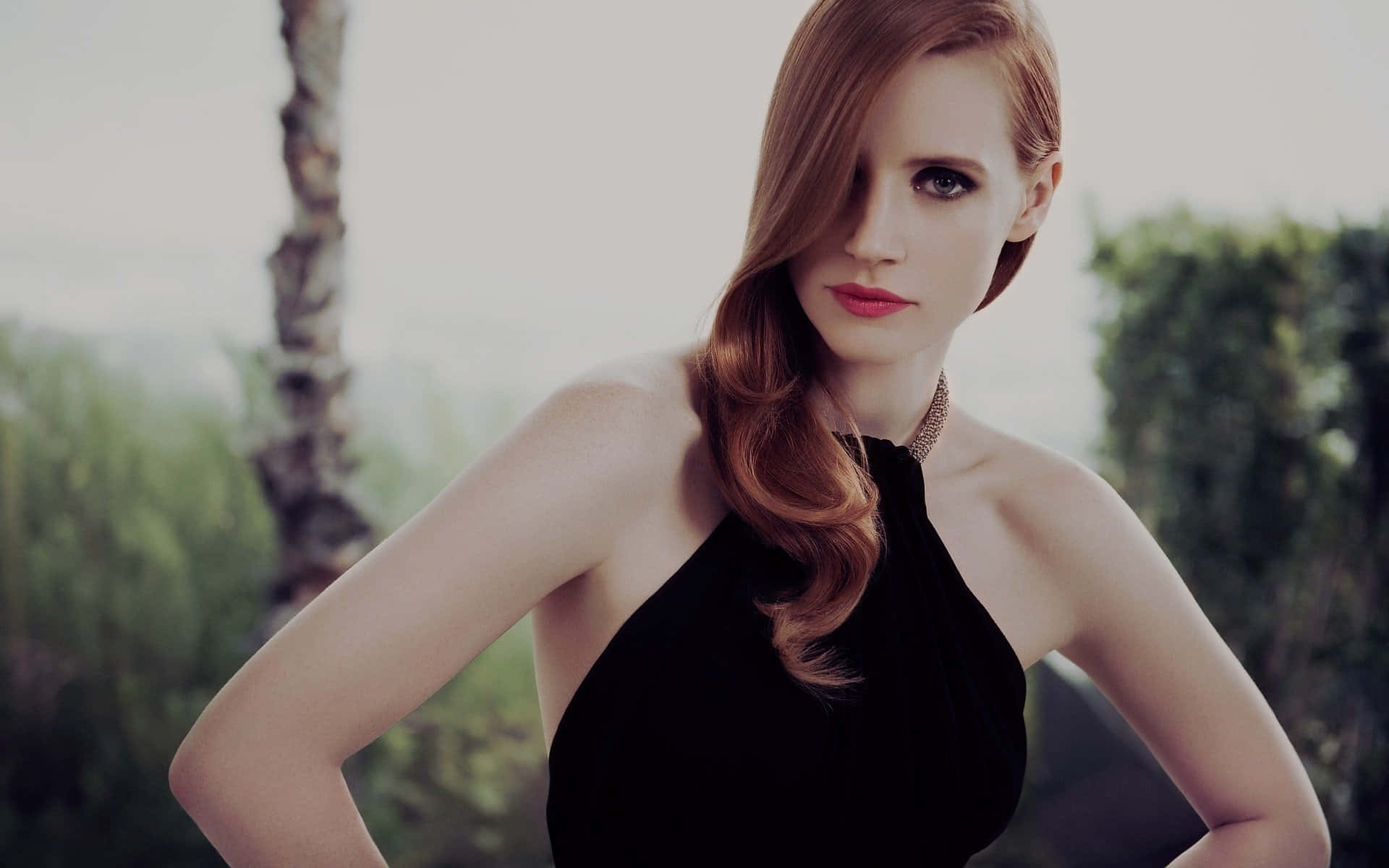 jessica chastain 桌布 壁紙 — 一位優雅的紅髮女性，塗著大膽的紅唇，穿著黑色掛脖連衣裙，背景是柔和的綠色戶外景色。