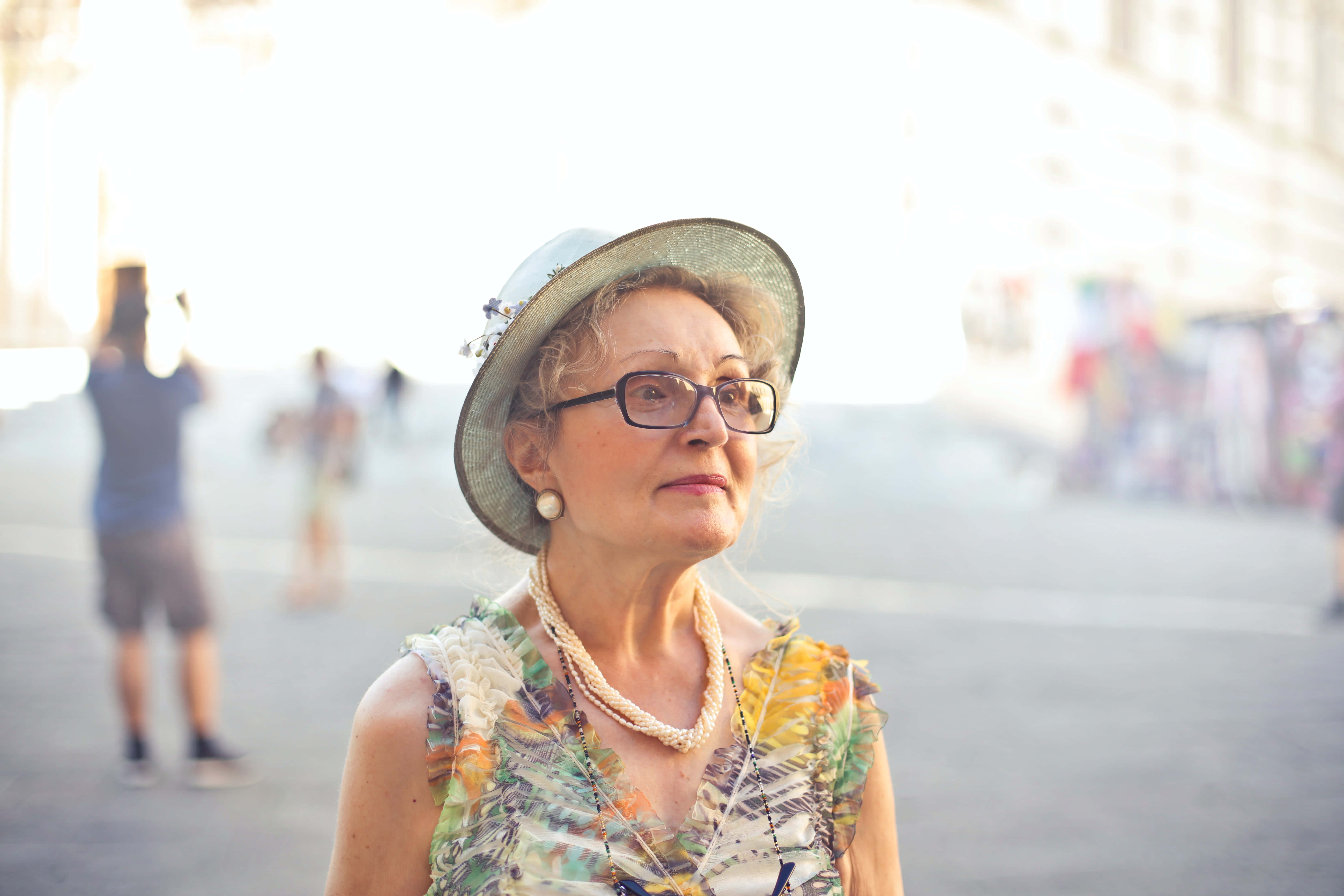 Summer Fashion 桌布 壁紙 — 一位優雅的老年女士穿著印花連衣裙和寬邊帽，在陽光明媚的城市廣場上若有所思地向上凝視。