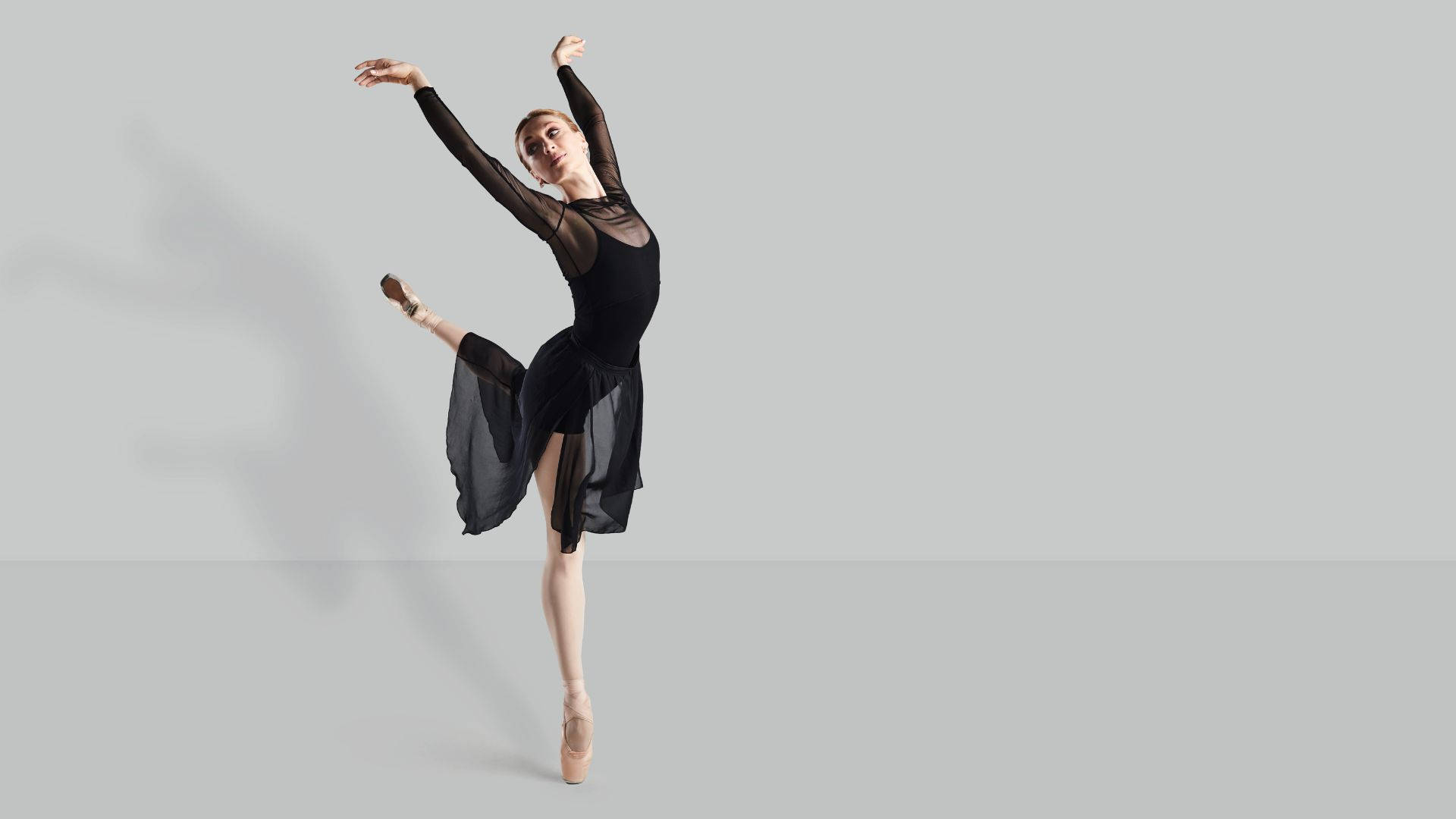ballerina 桌布 壁紙 — 一位優雅的芭蕾舞者身穿黑色緊身衣和飄逸裙子，在極簡灰色工作室背景下以足尖演繹優美的阿拉貝斯克姿勢。