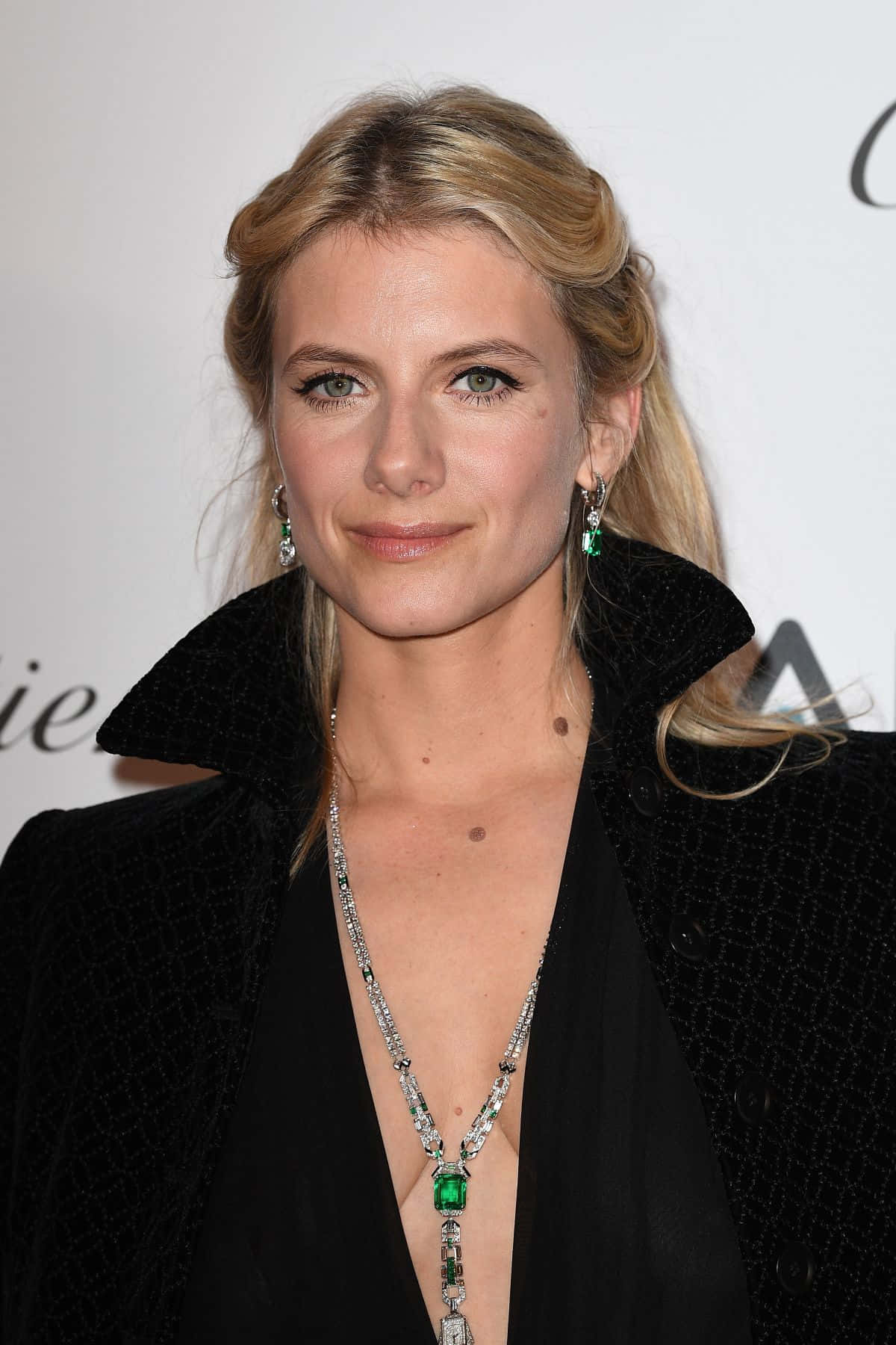 Mélanie Laurent 桌布 壁紙 — 一位優雅的金髮女士身穿黑色外套，在正式活動中佩戴一條引人注目的祖母綠和鑽石項鍊。