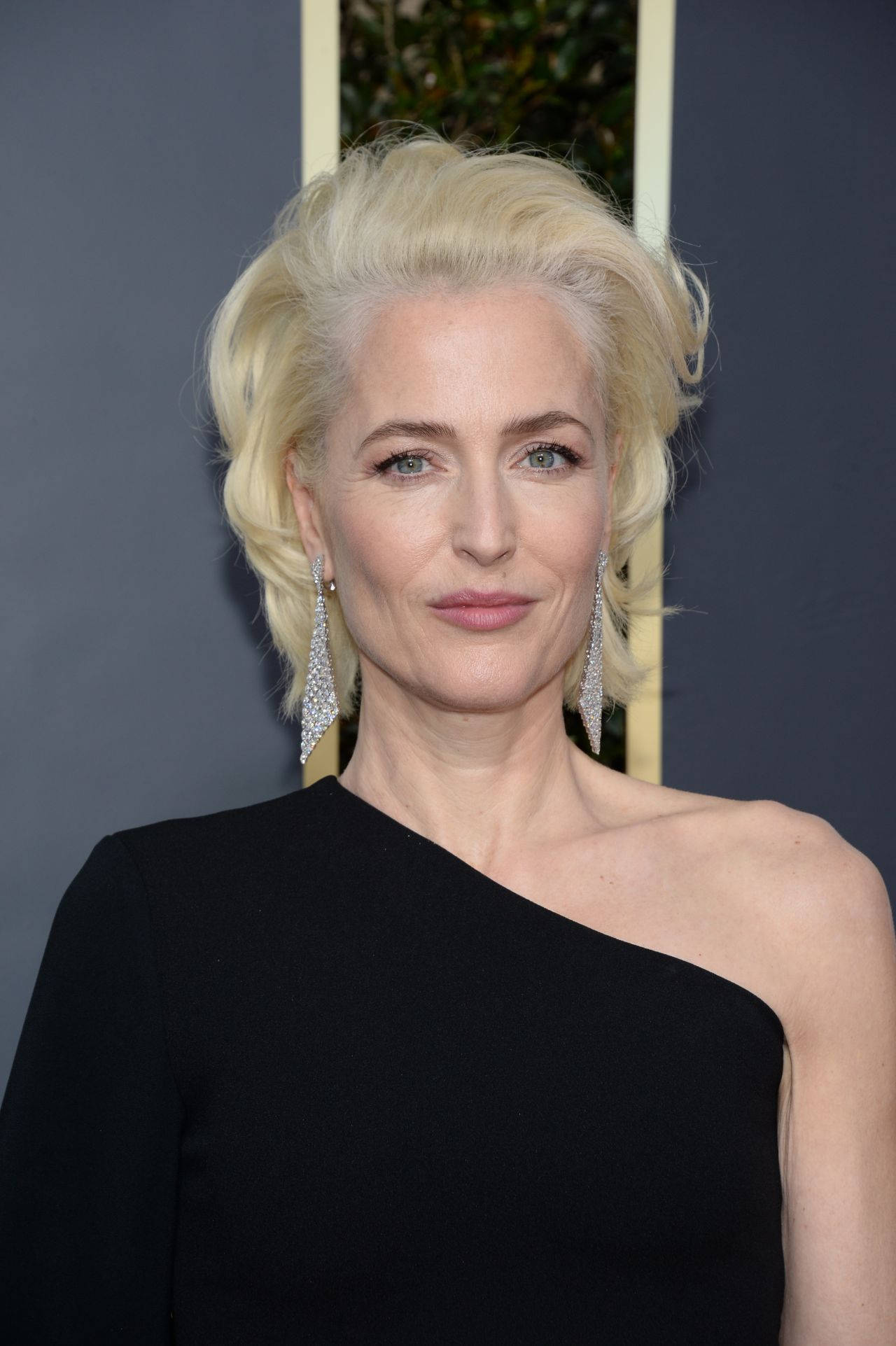 gillian anderson 桌布 壁紙 — 一位優雅的金髮女郎，戴著閃亮的耳環，身穿時尚的黑色單肩連衣裙，背景為深色。