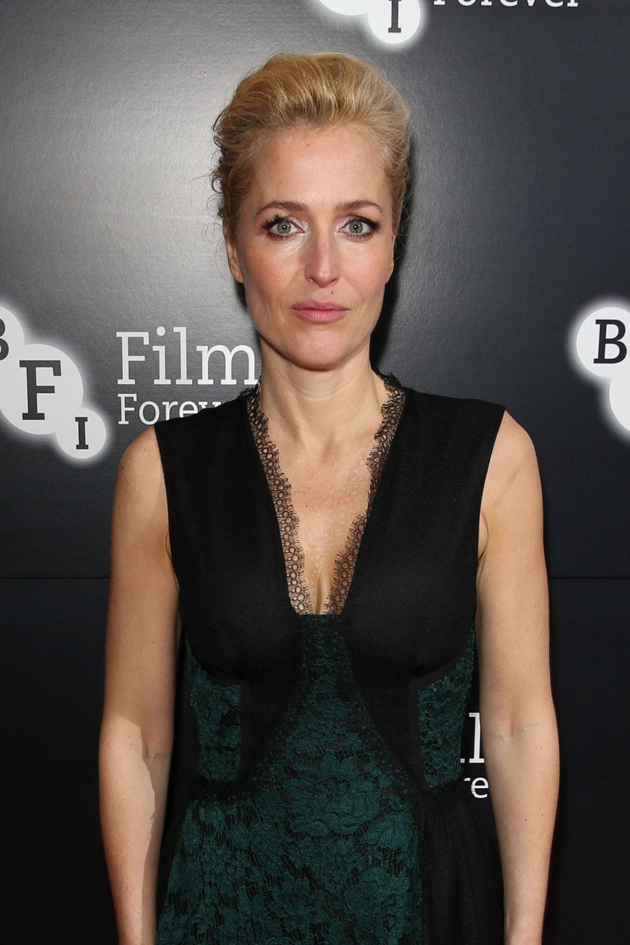 gillian anderson 桌布 壁紙 — 一位優雅的金髮女郎穿著黑色和綠色蕾絲連衣裙，站在帶有 'Film Forever' 標誌的深色背景前。
