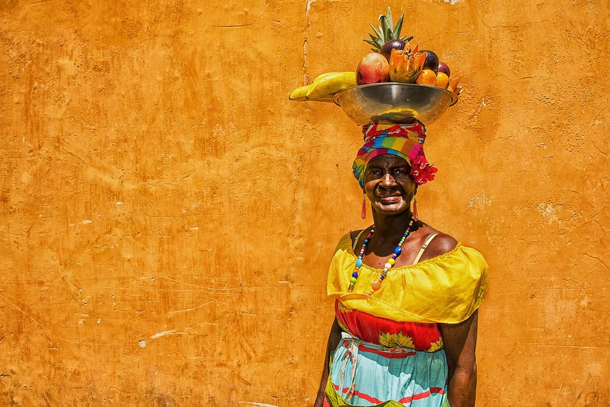 Colombia 桌布 壁紙 — 一位充滿活力的女士身穿色彩繽紛的傳統服飾，在紋理橙色的牆壁前，頭頂平衡一碗熱帶水果。