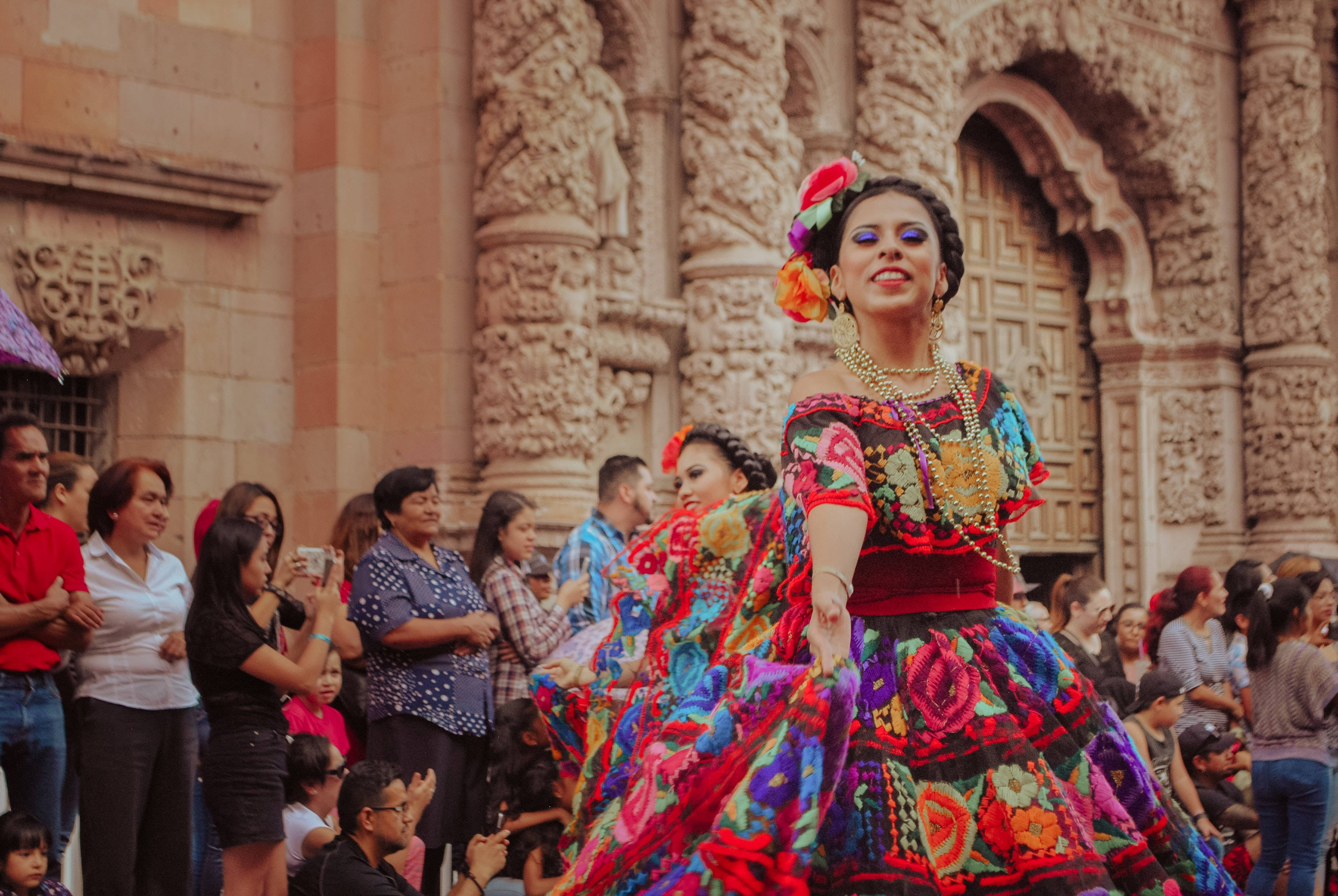 Mexican 桌布 壁紙 — 一位充滿活力的舞者穿著色彩繽紛的刺繡連衣裙，在華麗的石建築前為人群表演。
