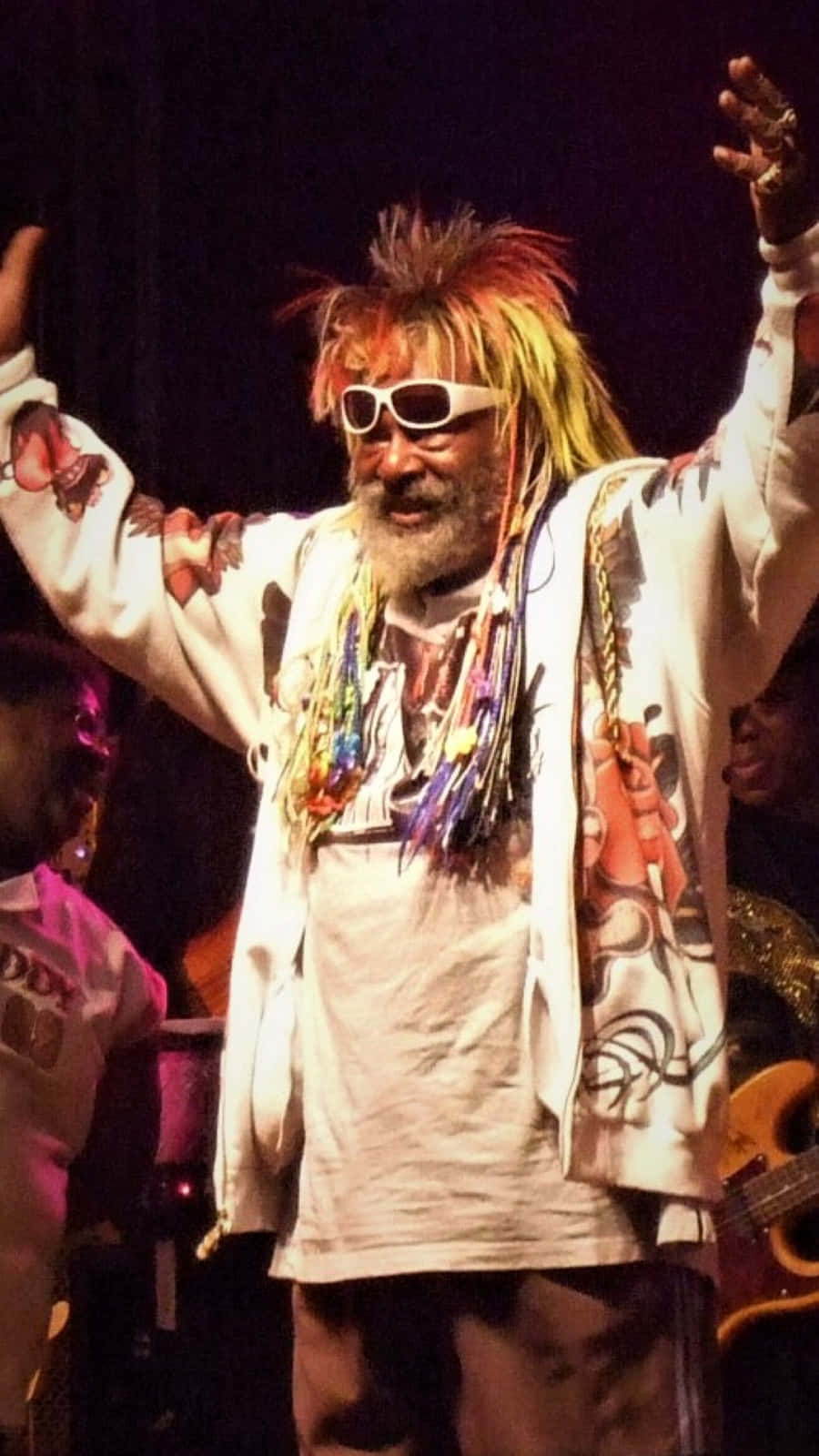 Famous 桌布 壁紙 — 一位充滿活力的表演者，擁有尖刺彩虹頭髮和白色太陽眼鏡，在明亮的舞台燈光下舉起雙臂。