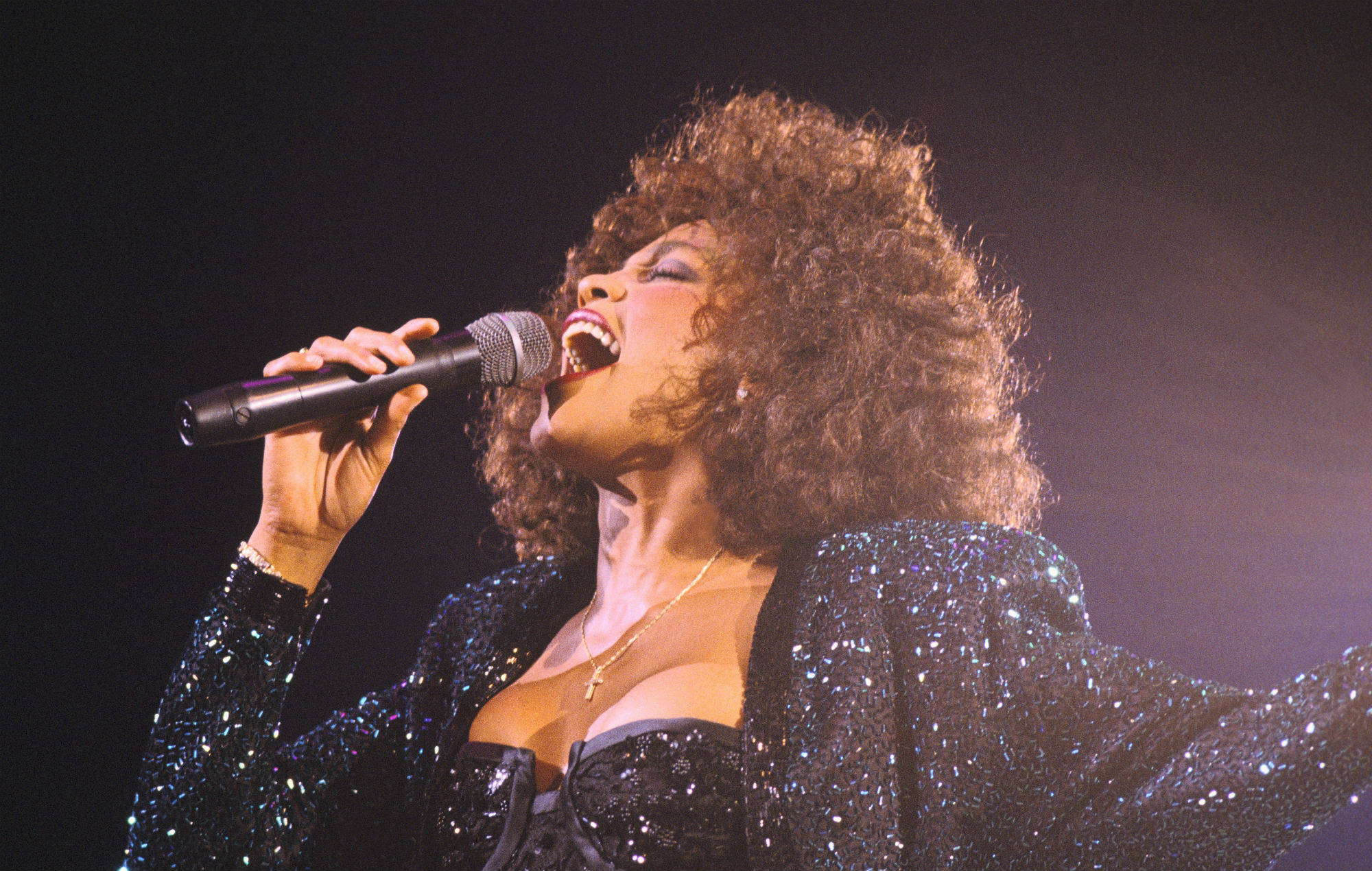 Whitney 桌布 壁紙 — 一位充滿活力的表演者，留著濃密的非洲式髮型，在舞台燈光下穿著閃亮的黑色服裝，熱情地對著麥克風唱歌。