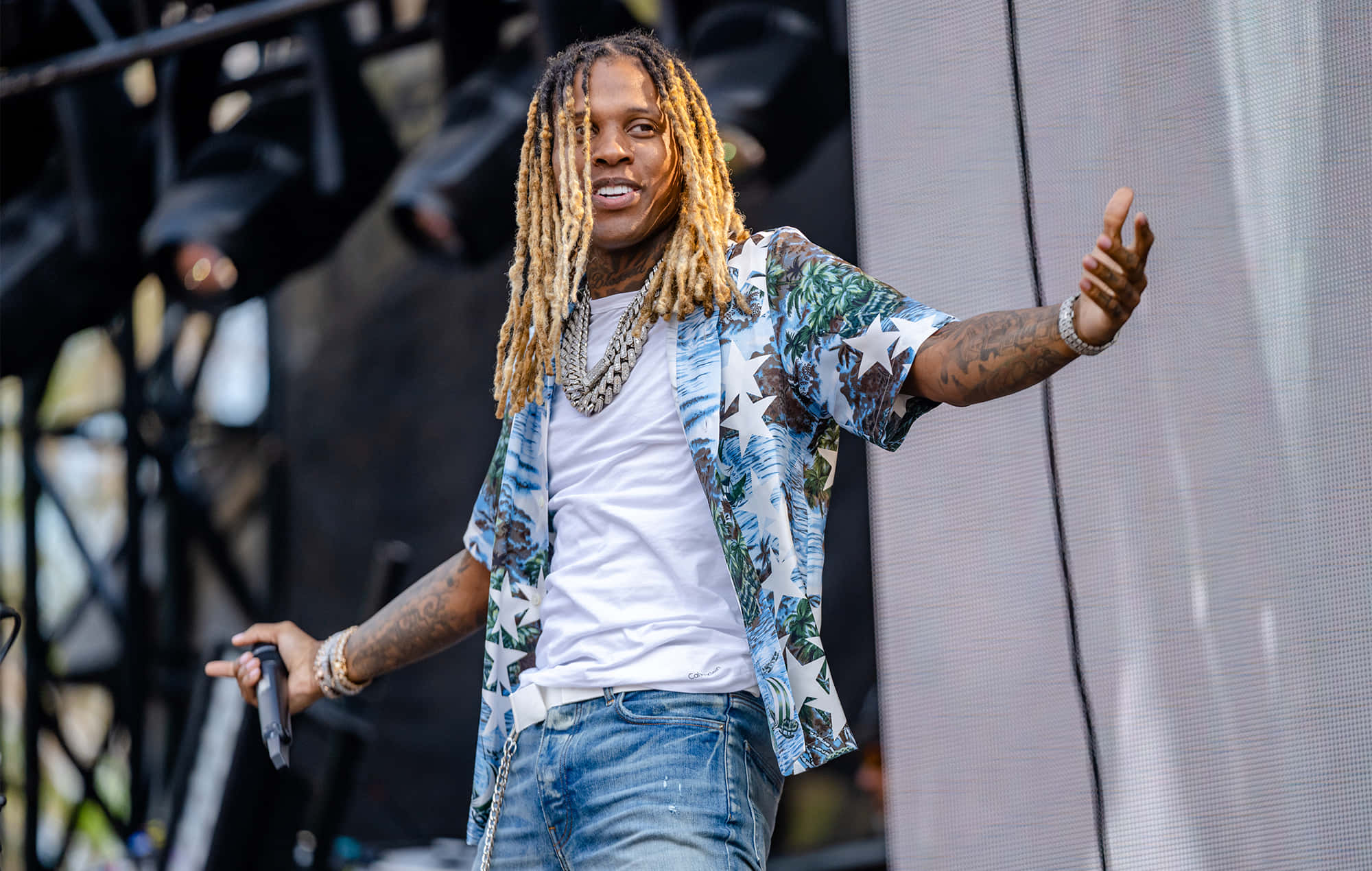 Lil Durk 桌布 壁紙 — 一位充滿活力的表演者，留著長辮子，身穿星星圖案襯衫，在陽光普照的戶外舞台上為人群注入能量。